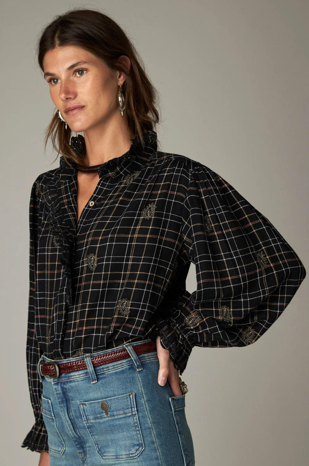 A woman wearing a black plaid Coco Blouse - Noir Plaid with a ruffled collar and cuffs and small embroidered details.