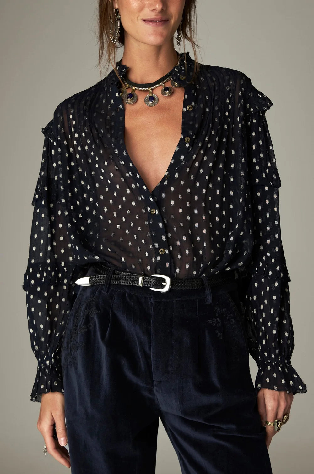 Maison Hotel Simona Blouse in Sienna navy, a metallic jacquard top with white polka dots, ruffle shoulders, and a V-neckline, worn with dark trousers and a thin braided belt.