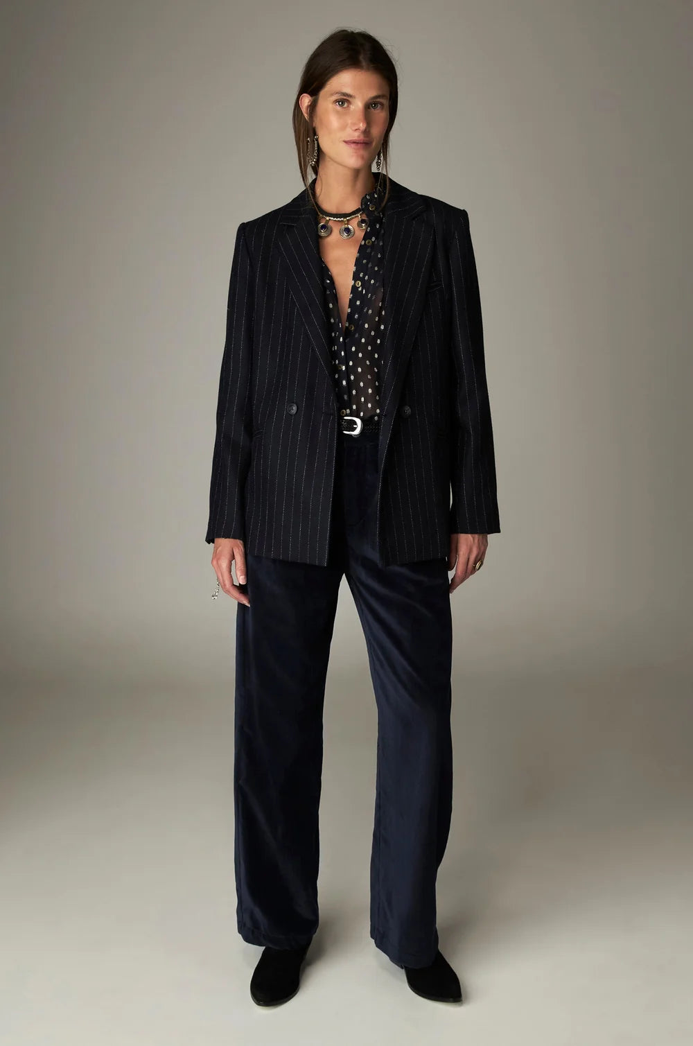A person wearing a black pinstripe blazer over a sheer, polka-dot metallic jacquard Simona Blouse - Sienna navy and dark velvet wide-leg trousers.