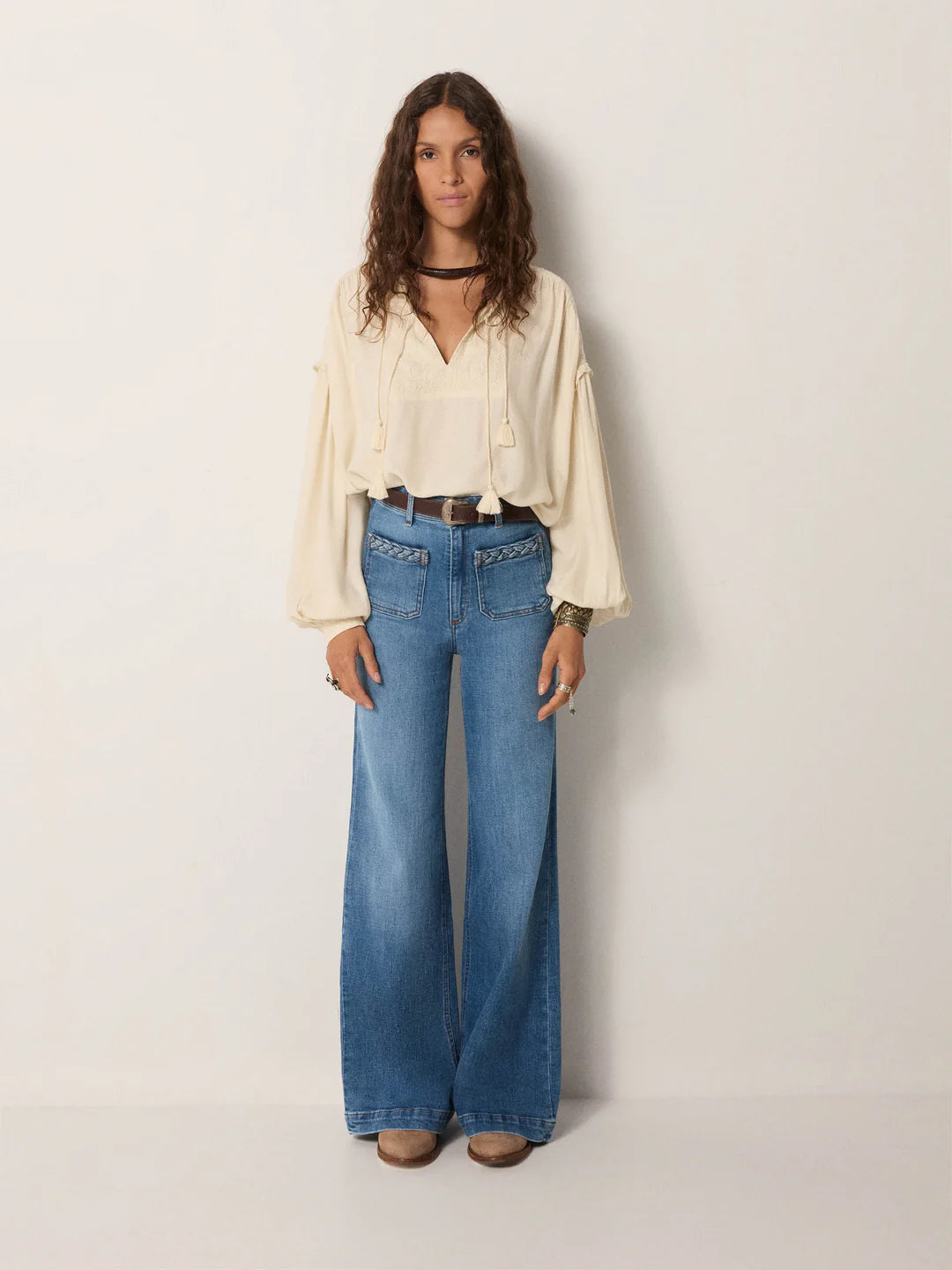 A woman stands against a plain wall wearing a cream blouse, tan shoes, and Maison Hotel's Giuletta Trousers in light blue with a brown belt.