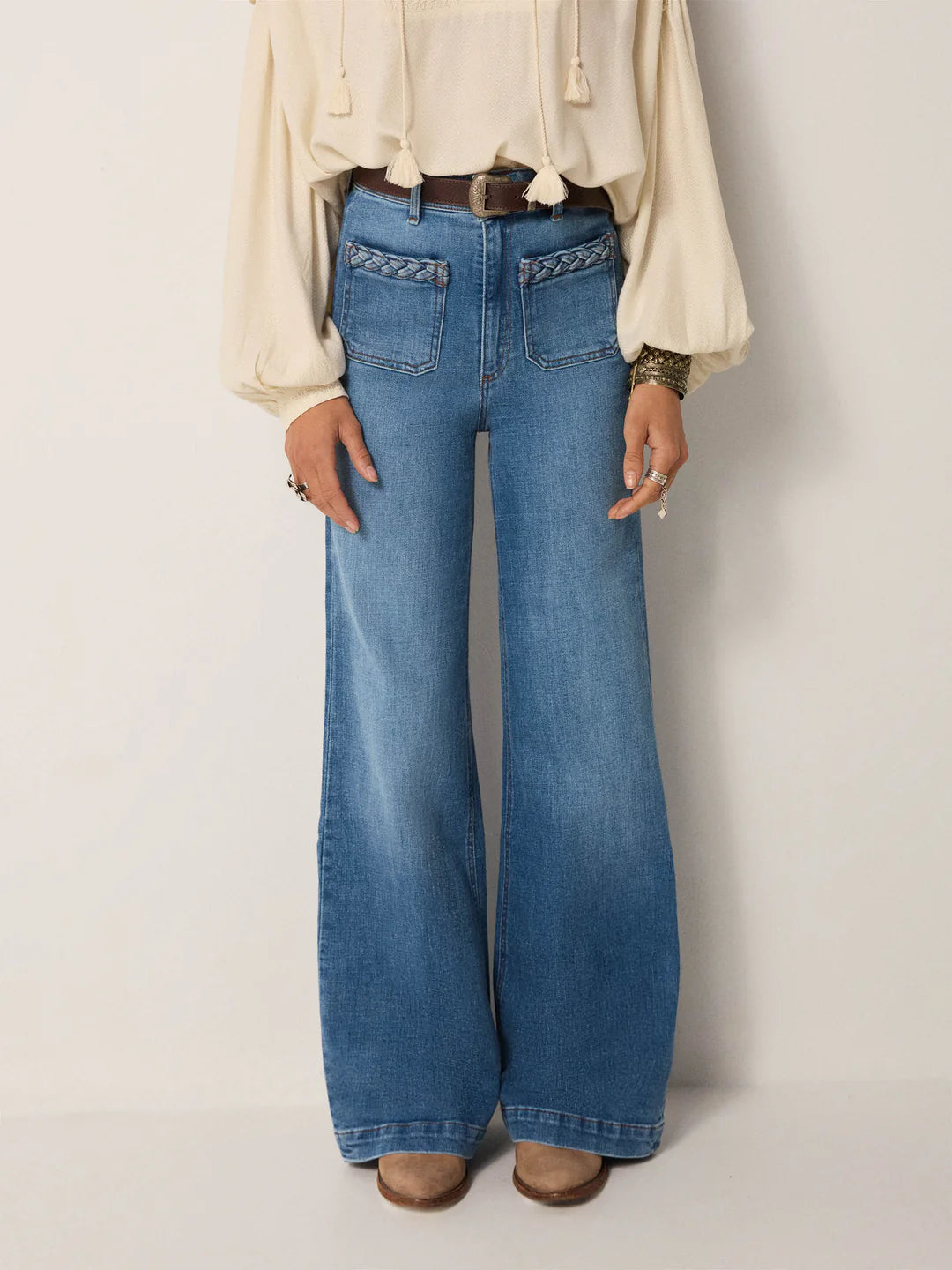 A person stands against a plain background wearing Maison Hotel’s Giuletta Trousers in light blue, featuring a high rise, front pockets, and a braided waistband, paired with a cream blouse and brown shoes.