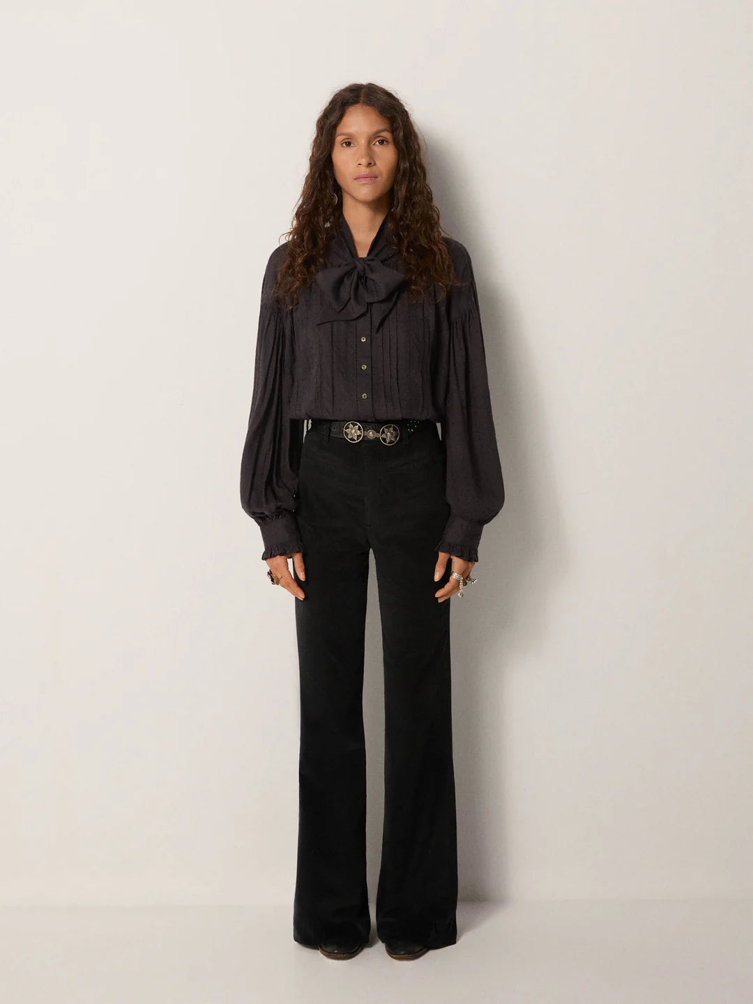 A woman stands against a plain wall wearing the Lorraine Blouse in Twilight by Maison Hotel, paired with black high-waisted pants and a decorative belt.