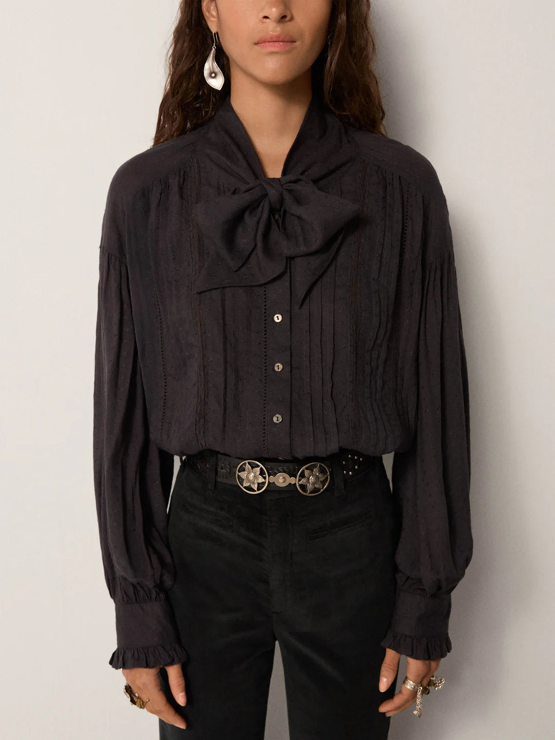 Wearing the Maison Hotel Lorraine Blouse - Twilight with a large neck bow, black high-waisted pants, a silver belt, and a single dangling earring, the person stands against a plain background.