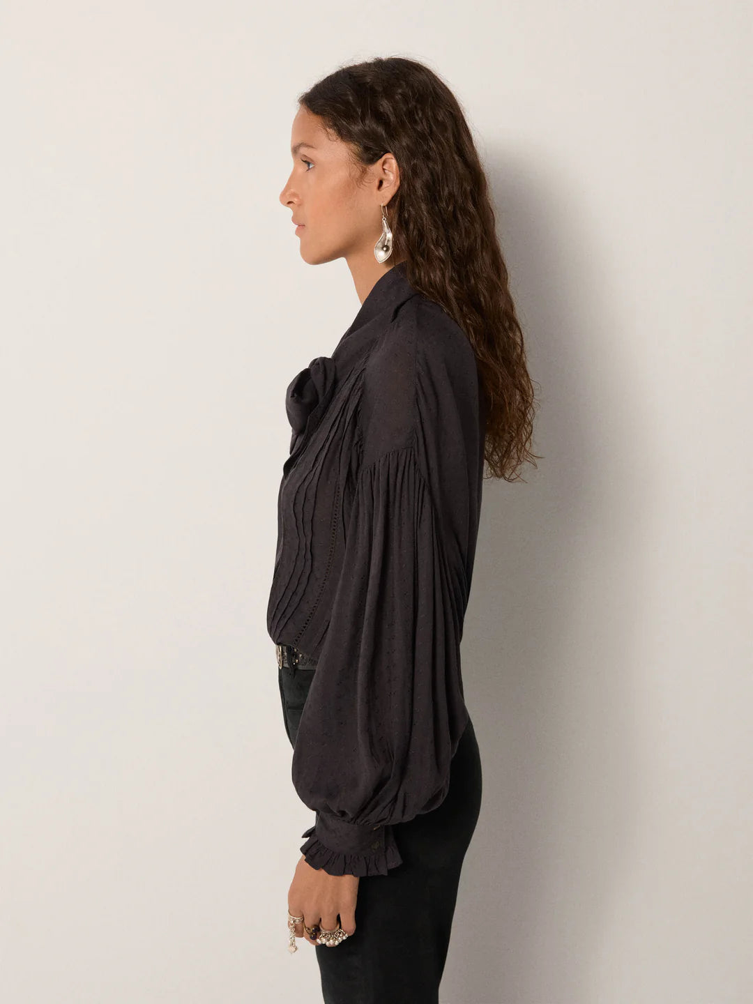 Woman with long curly hair wears the Maison Hotel Lorraine Blouse in Twilight, styled with earrings and rings, standing in profile against a plain light background.