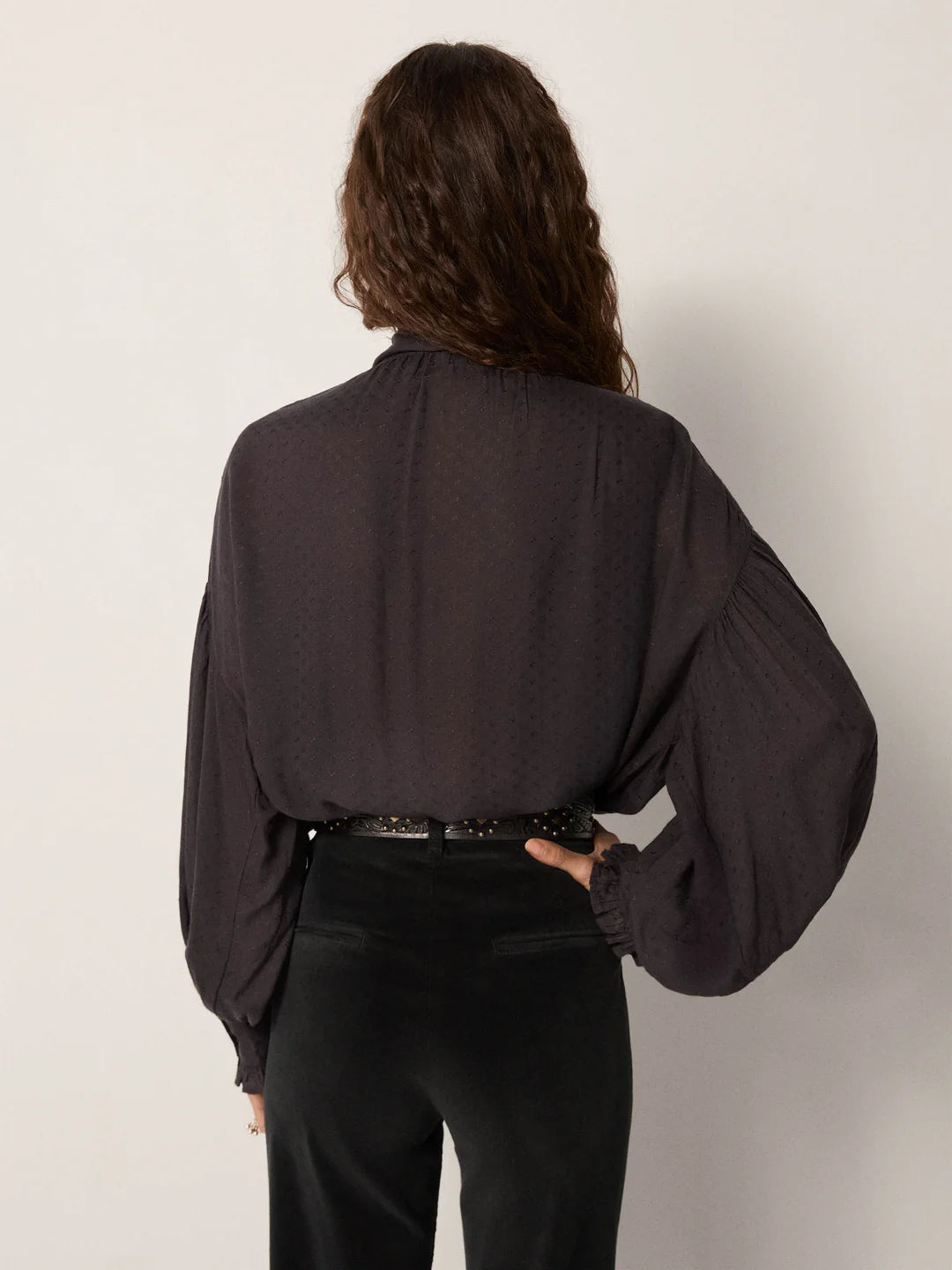 A woman with long wavy hair is shown from the back, wearing the Maison Hotel Lorraine Blouse in Twilight with billowy sleeves, tucked into high-waisted black pants and cinched with a black belt.