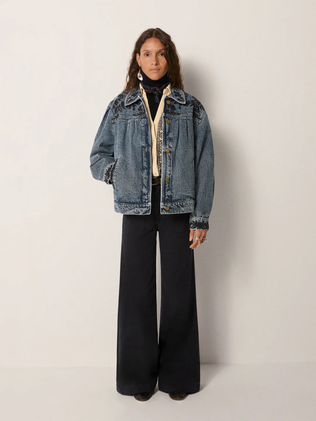 A person stands against a plain background wearing the Maison Hotel Wesley Denim Jacket – Western Blue with subtle floral embroidery, layered over a turtleneck and wide-leg black pants, hands tucked in the jacket pockets.