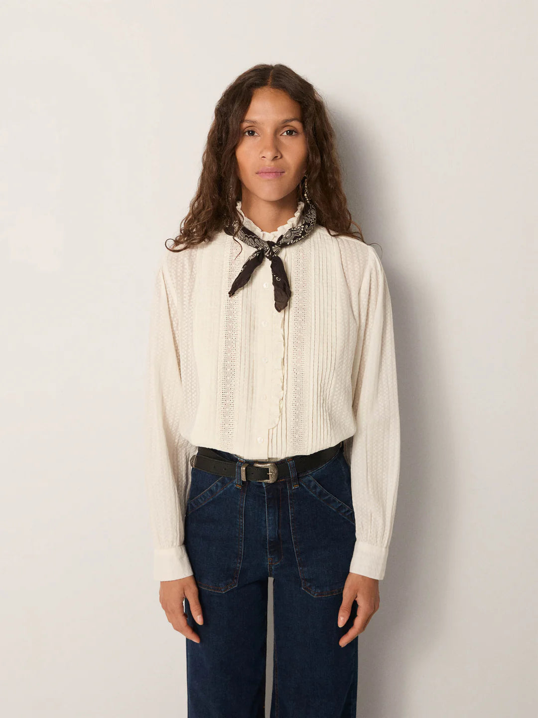 A woman with long curly hair faces forward, wearing the Maison Hotel Bella Blouse in white, paired with dark blue jeans, a black belt, and a black patterned neck scarf.