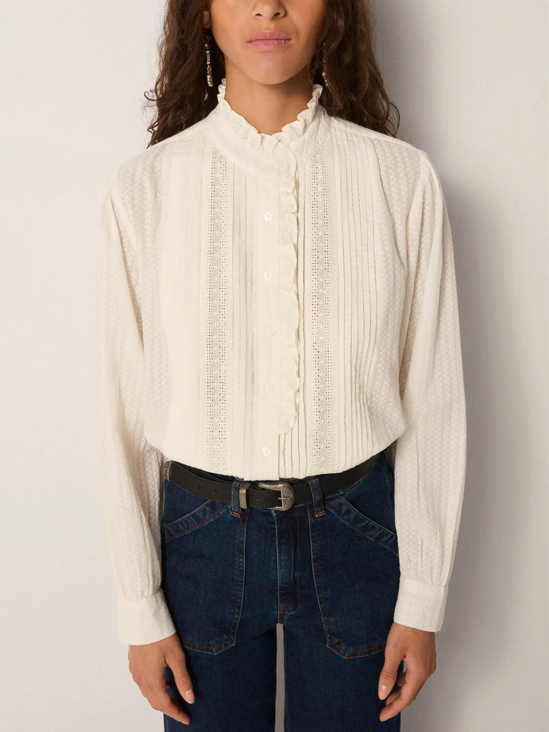 A person wearing the Maison Hotel Bella Blouse in white, featuring a ruffle collar and delicate details, styled with dark blue jeans and a black belt.