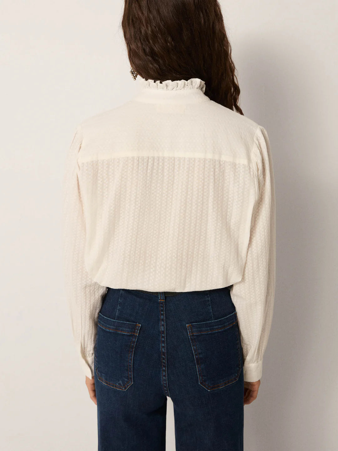 A person with long brown hair wears the Maison Hotel Bella Blouse in white, a long-sleeve lace top with a ruffle collar, paired with blue jeans and seen from the back against a plain background.