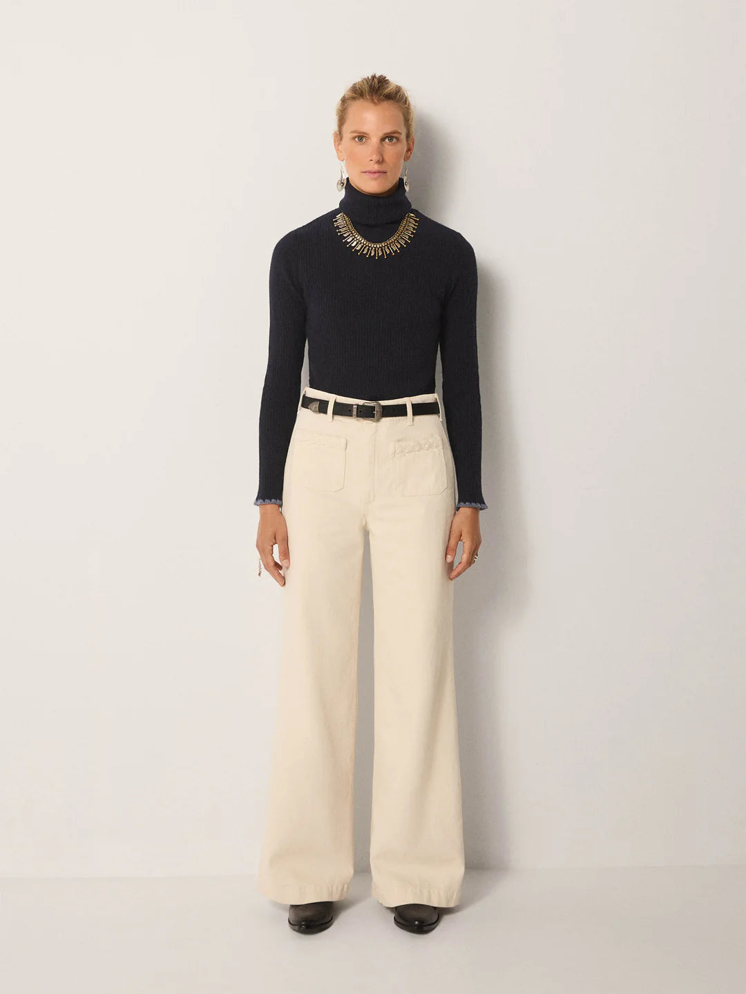 A person stands against a plain background wearing a navy Dolcevita Sweater by Maison Hotel, cream wide-leg pants, a statement necklace, a belt, and black shoes.