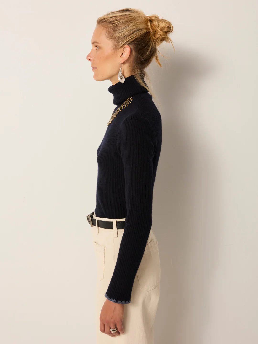 A person stands in profile against a plain background, wearing the Maison Hotel Dolcevita Sweater in navy, paired with light-colored belted pants and earrings.