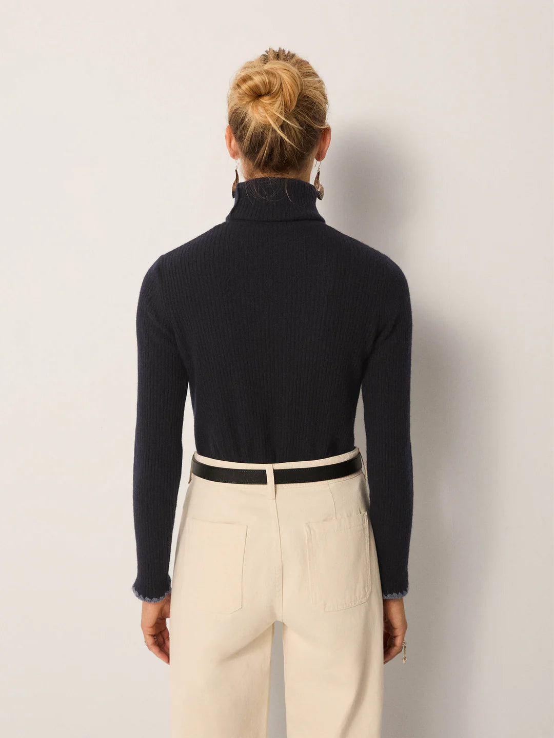 A woman stands facing away, wearing a navy Dolcevita Sweater by Maison Hotel and high-waisted cream pants with a black belt. Her hair is styled in a neat bun.