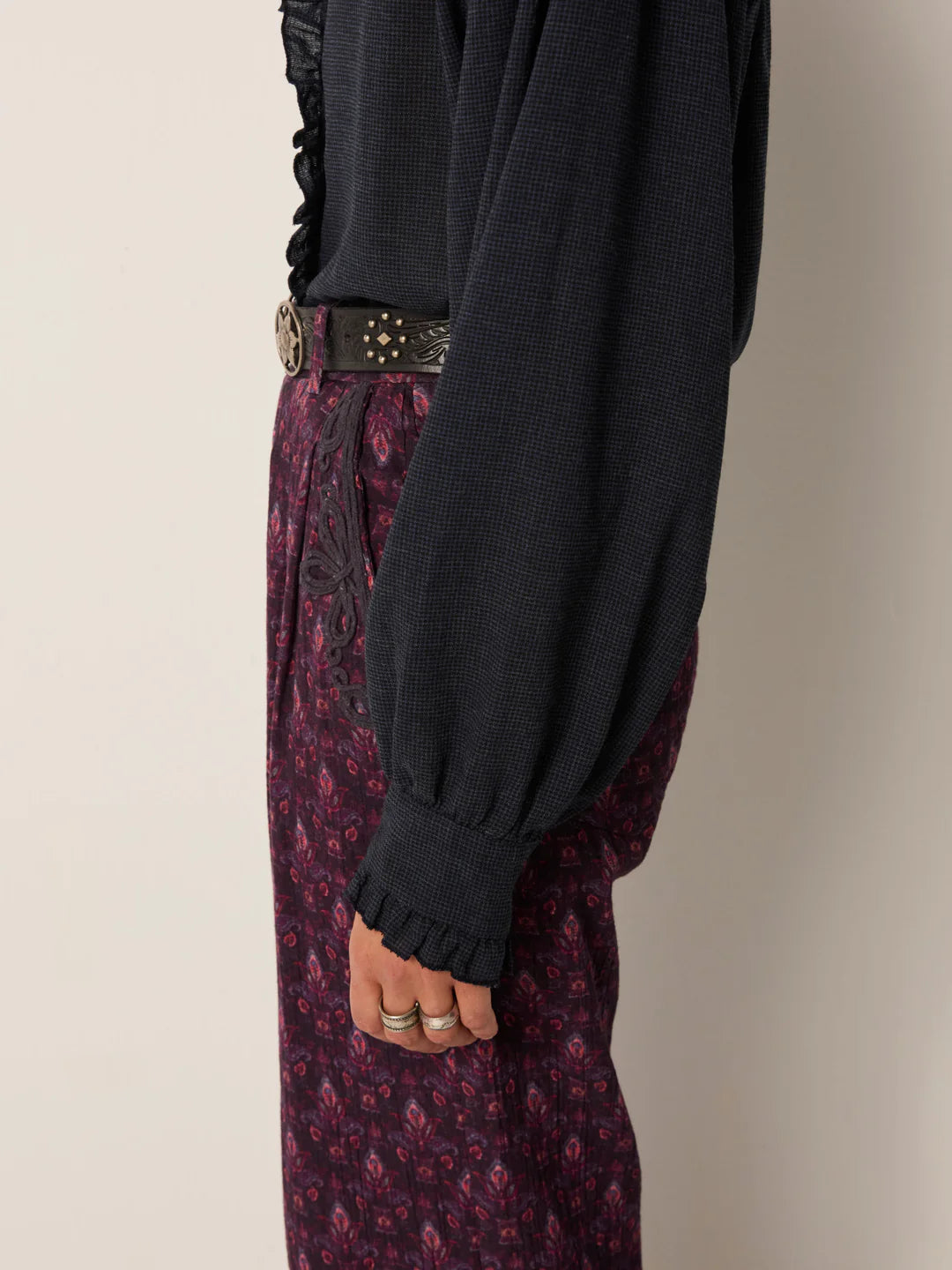 A person stands sideways wearing the Maison Hotel Celeste Blouse in Eclipse Blue with puffed sleeves and ruffle trim, paired with a patterned belt and high-waisted maroon patterned pants. Three rings are visible on their left hand.