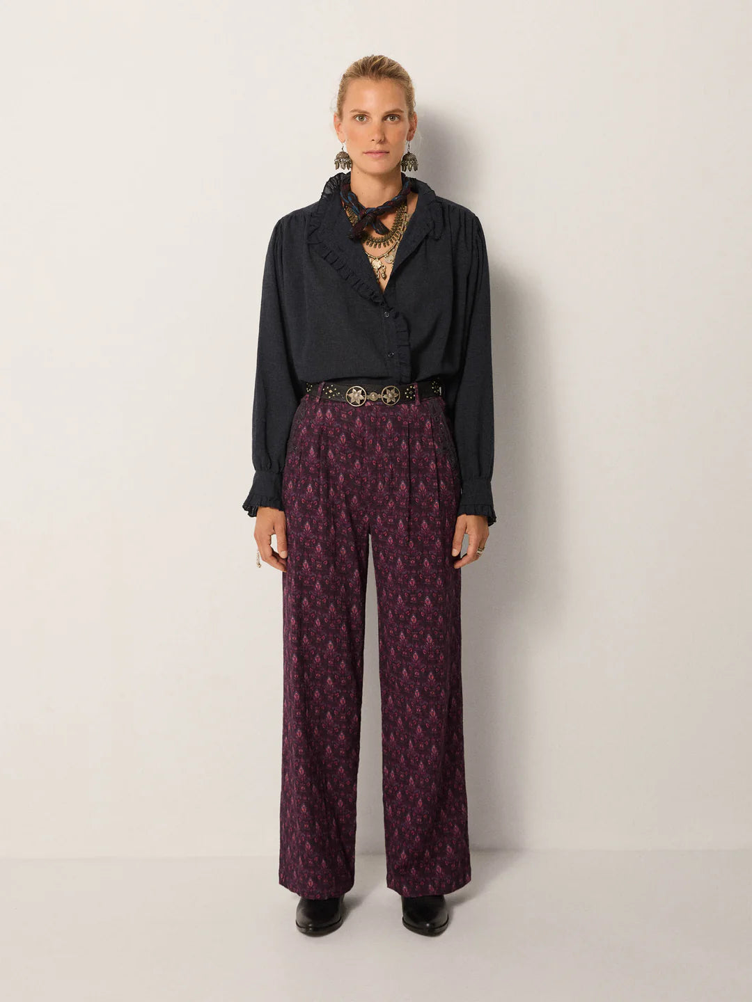 A woman stands against a plain wall wearing the Maison Hotel Celeste Blouse in Eclipse Blue, paired with patterned high-waisted wide-leg pants, a wide belt, black boots, and layered necklaces.