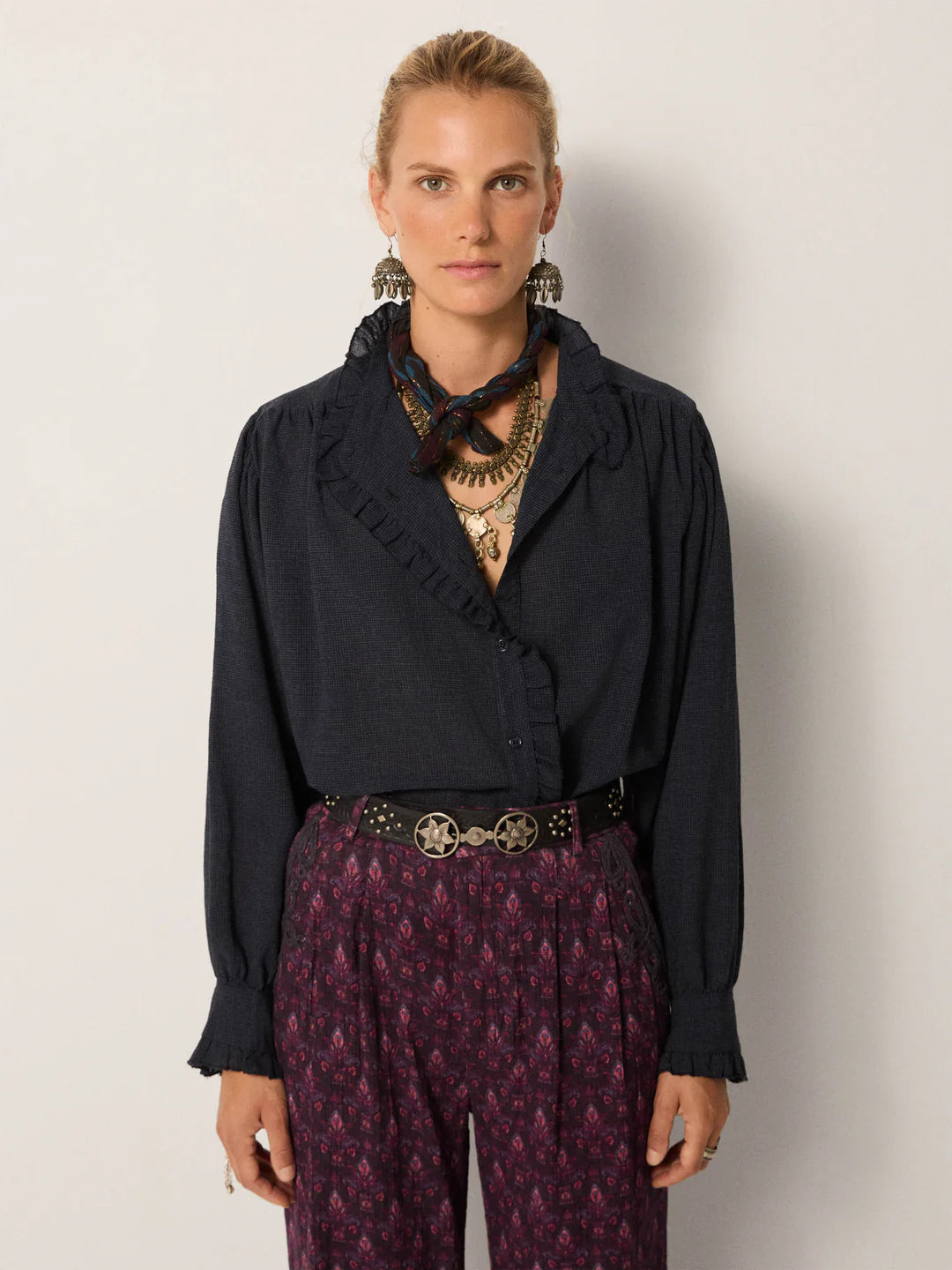 A woman stands against a plain background wearing the Maison Hotel Celeste Blouse in Eclipse Blue, paired with patterned pants, layered necklaces, earrings, a printed scarf, and a studded belt.