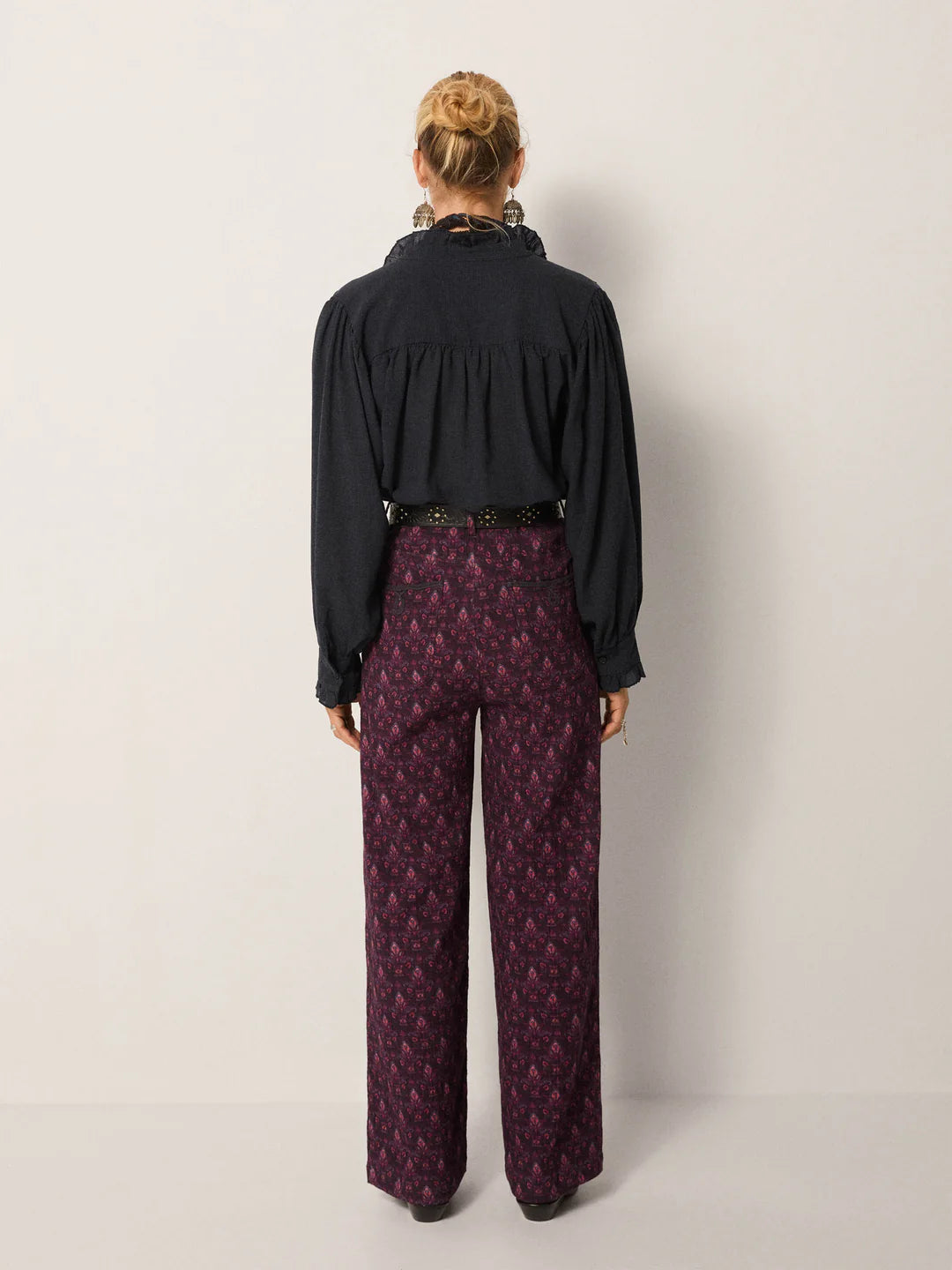 A person faces away from the camera, wearing the Maison Hotel Celeste Blouse in Eclipse Blue with subtle ruffle trim and high-waisted patterned maroon pants, standing against a plain light-colored wall.