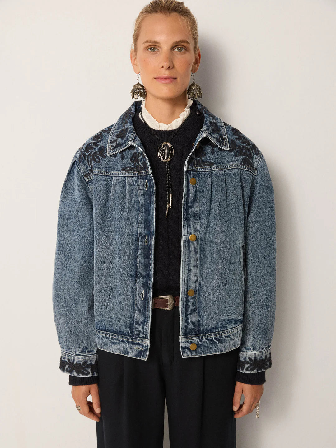 A woman stands against a plain light background wearing the Maison Hotel Wesley Denim Jacket in Western Blue over a dark sweater and black pants, featuring subtle floral embroidery.