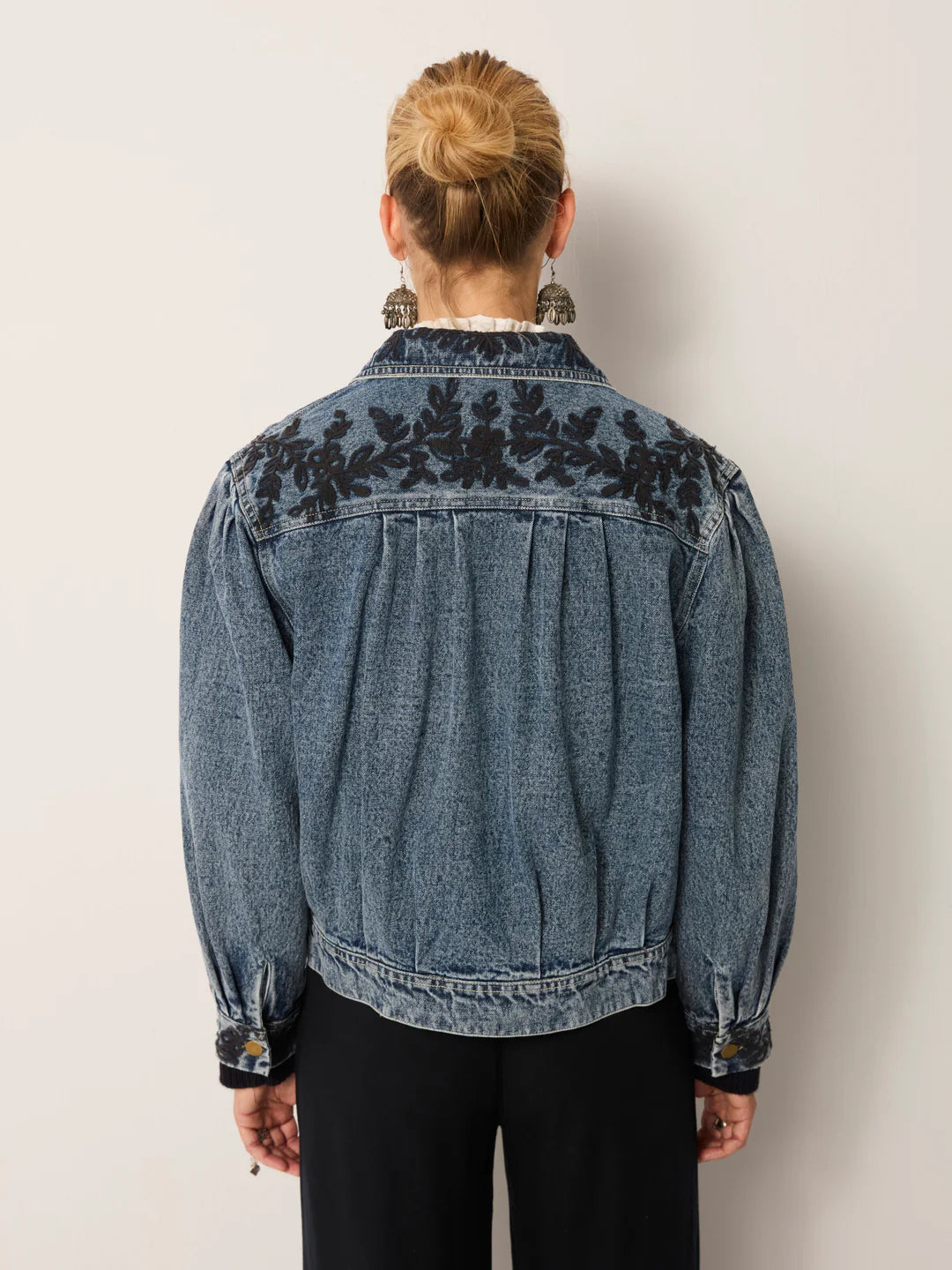 A person faces away in the Maison Hotel Wesley Denim Jacket in Western Blue, featuring black floral embroidery on the shoulders and cuffs, paired with black pants against a plain light background.