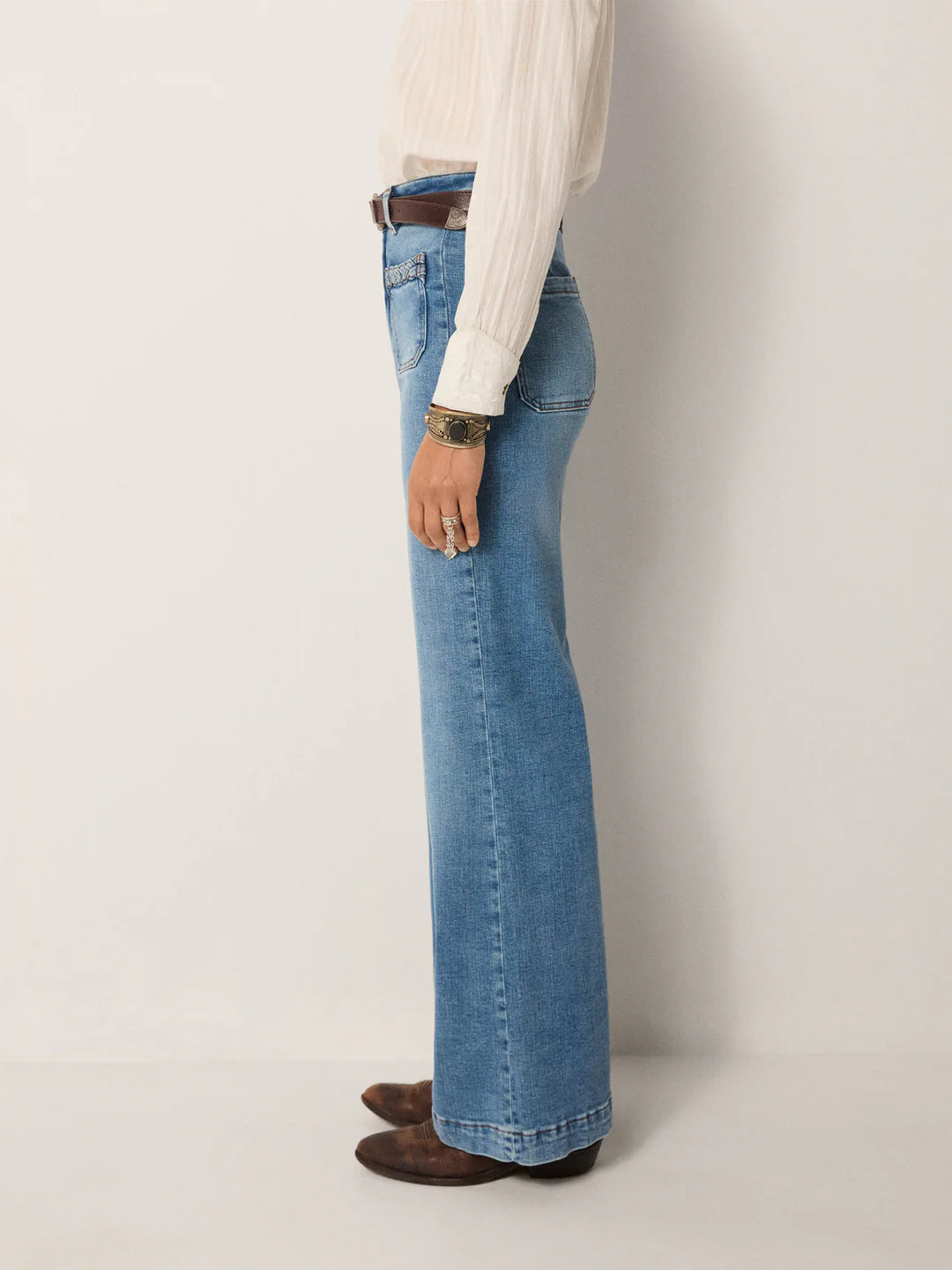 A person stands in profile against a plain background, wearing a white blouse and Maison Hotel’s Giuletta Trousers in light blue, styled with a brown belt and brown shoes.