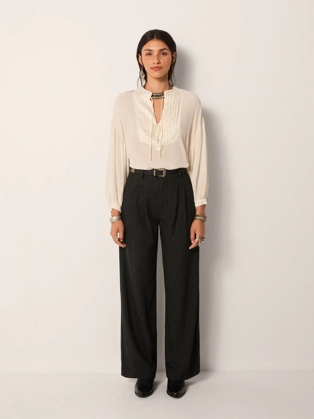 Person stands against a plain background, wearing the Maison Hotel Giovanni Blouse - Ivory with a pleated bib front and ruffled collar, paired with black belted wide-leg trousers and black shoes.