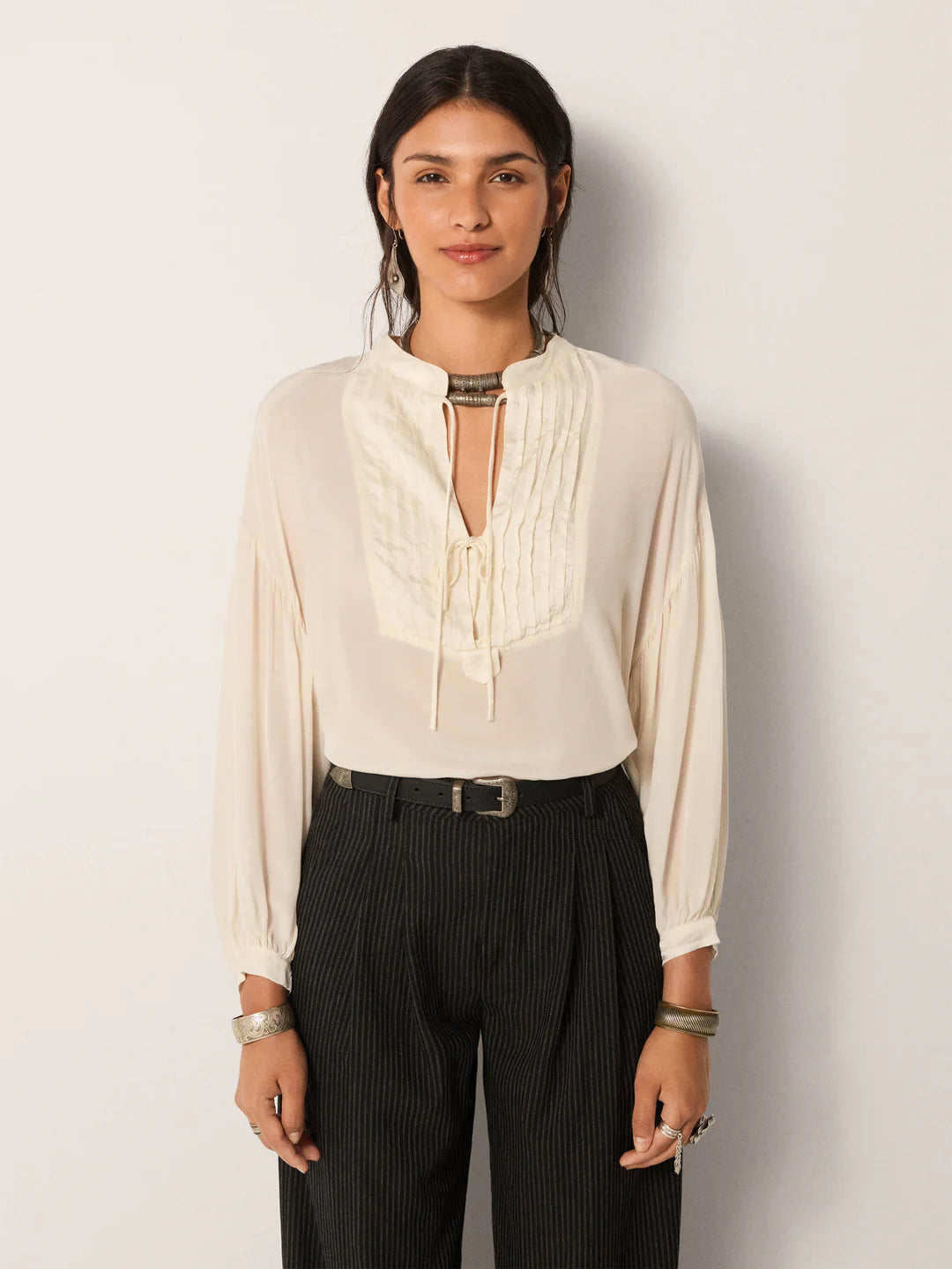 A person stands against a plain light background, wearing the Maison Hotel Giovanni Blouse in Ivory with black pinstripe trousers, accessorized with jewelry and a belt.