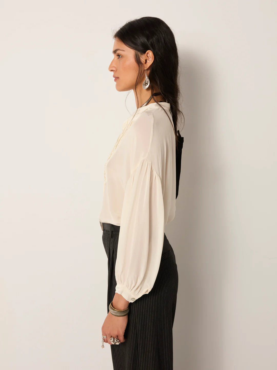 A woman stands in profile against a plain background, wearing the Maison Hotel Giovanni Blouse in Ivory with pleated bib front and high-waisted dark striped pants, styled with minimal jewelry for a vintage-inspired look.