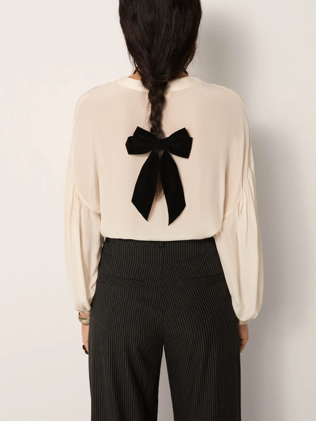 A person with a long braid, shown from behind, wears the vintage-inspired Giovanni Blouse in Ivory by Maison Hotel, featuring a large black bow on the back and dark pinstriped pants.