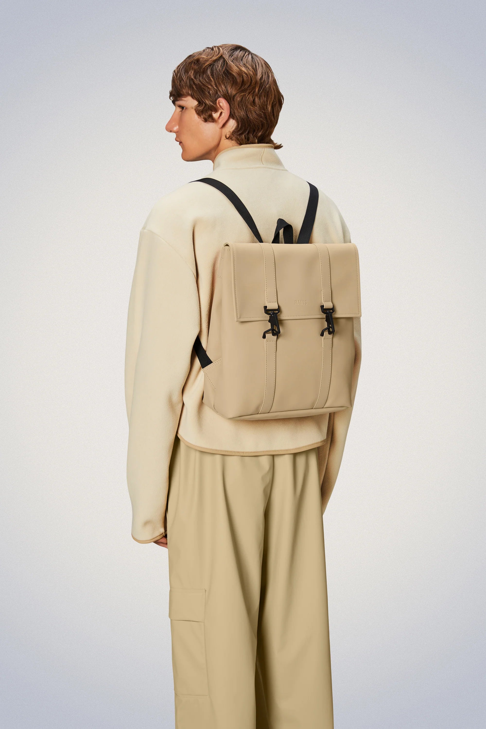 The back of a man wearing tan pants and a beige Rains MSN Bag Mini, featuring the classic school backpack design.