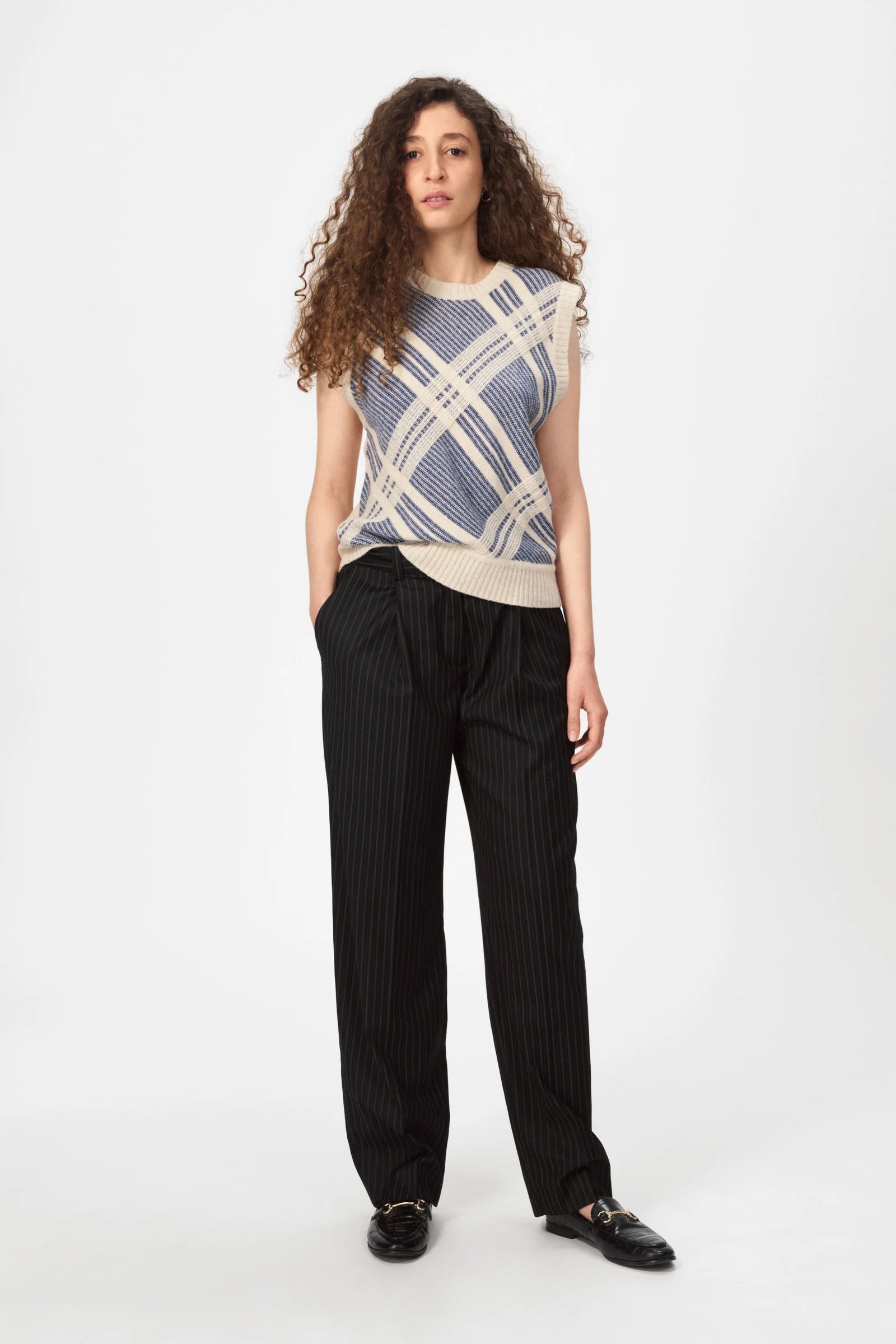 A person with long curly hair wears the Nué Notes Malik Vest - Egret Marine, paired with black pinstriped trousers and black shoes, standing against a plain white background.