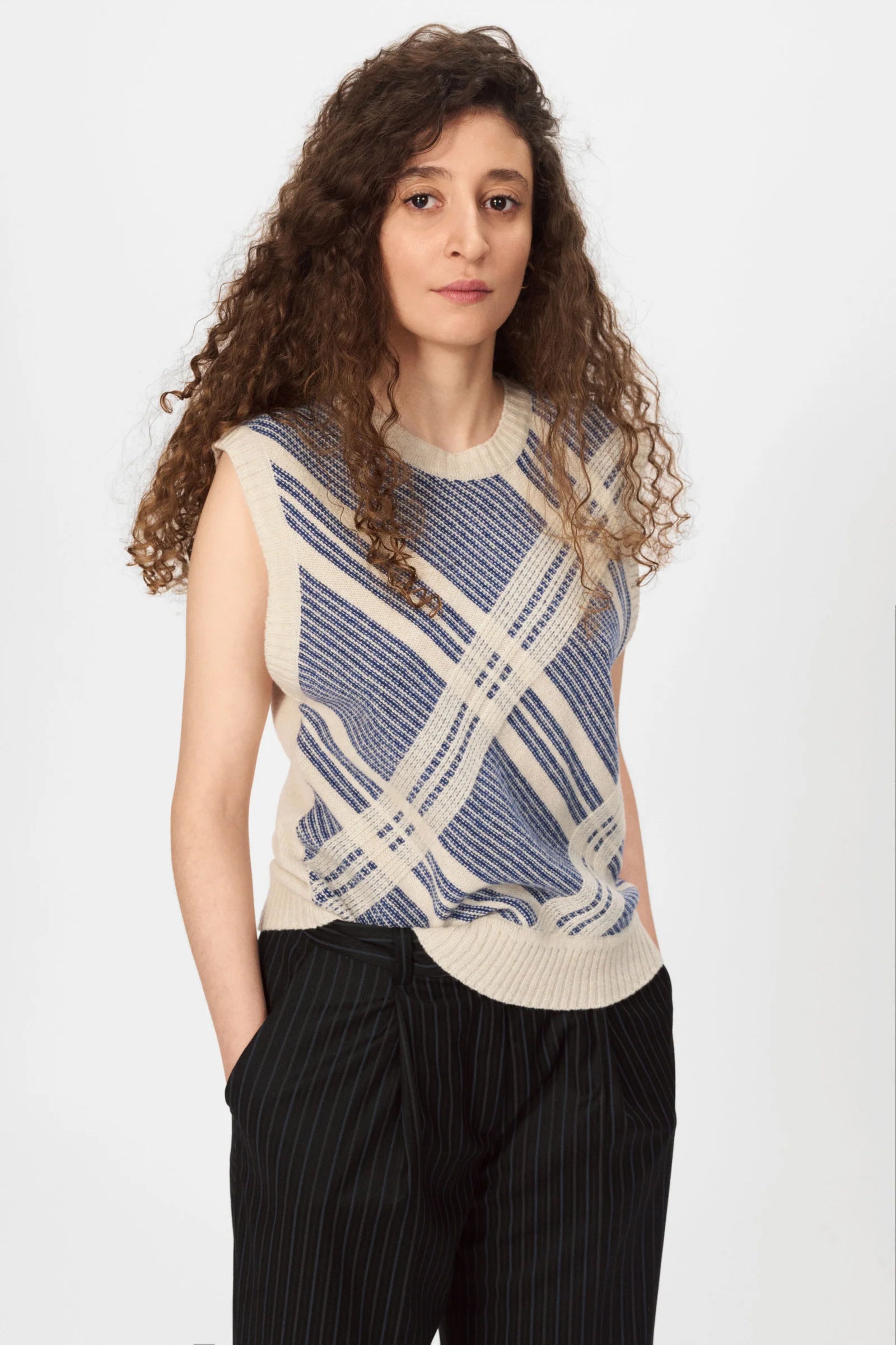 A person with long curly hair wears the Nué Notes Malik Vest - Egret Marine, paired with black pinstripe trousers, standing against a plain white background.