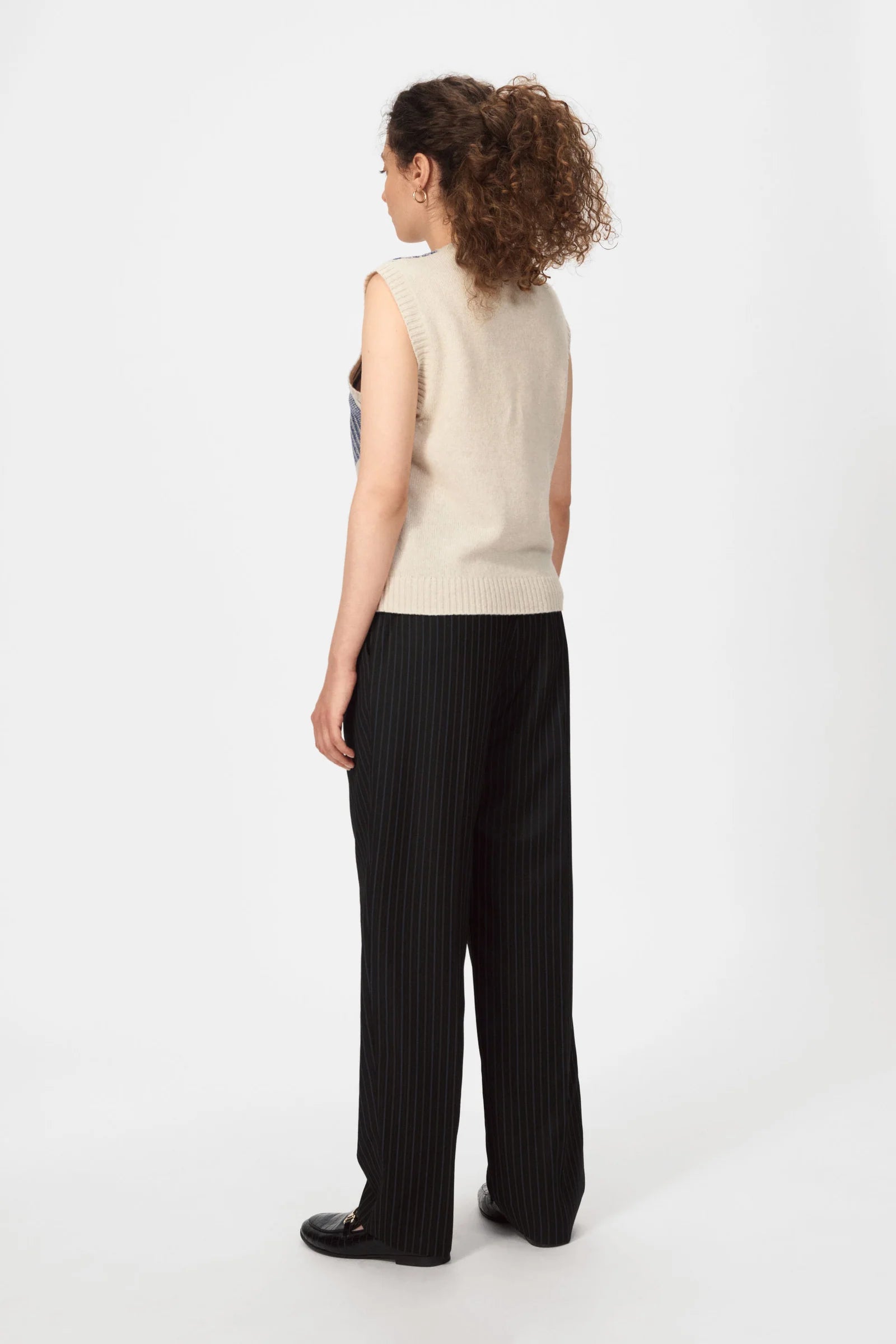 A woman with curly hair faces away, wearing a Nué Notes Malik Vest in Egret Marine, paired with black pinstripe pants and black shoes, set against a plain white background.