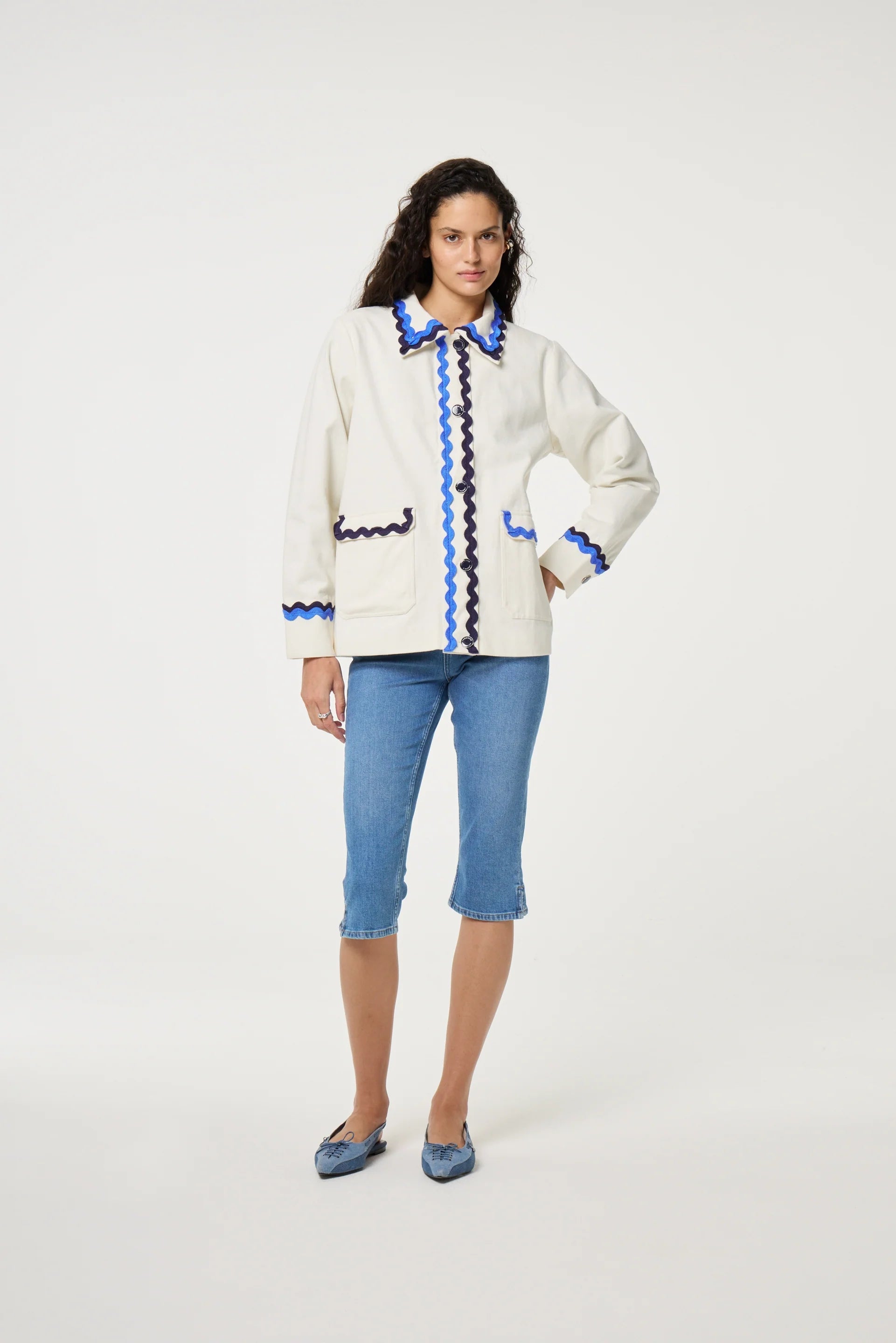 A woman wears the Fabienne Chapot Melodie Jacket in Warm White, featuring blue trim, styled with blue knee-length jeans and blue shoes against a plain white background, highlighting this versatile piece.