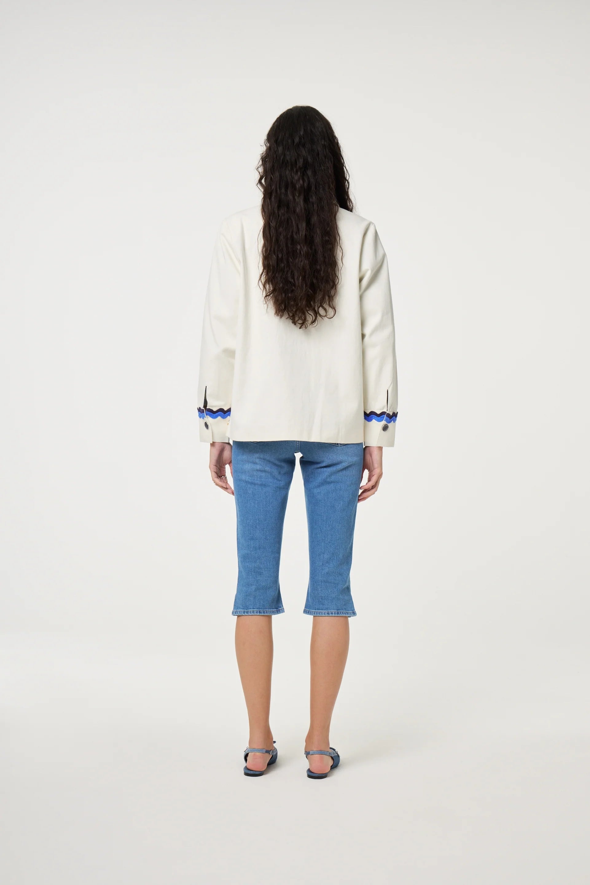 A person facing away wears the Fabienne Chapot Melodie Jacket - Warm White with blue trim, knee-length blue denim pants, and sandals against a white background—a versatile piece for any season.