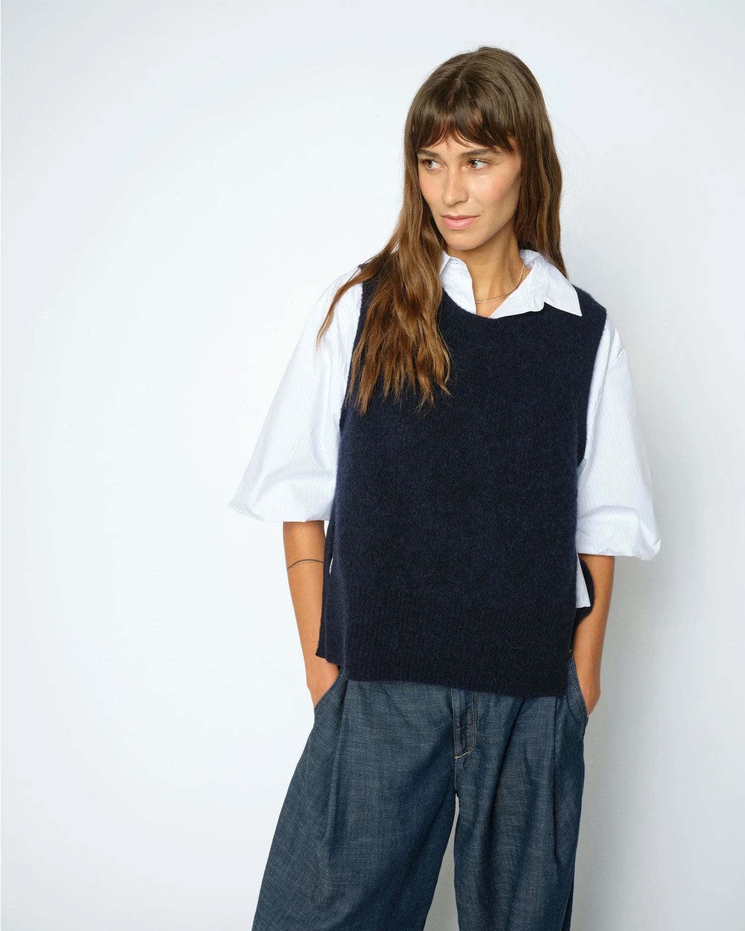 A woman with long brown hair stands against a plain light background with her hands in her pockets, wearing the MOS MOSH Thora Tie Knit Vest in navy over a white collared shirt and wide-leg blue pants.