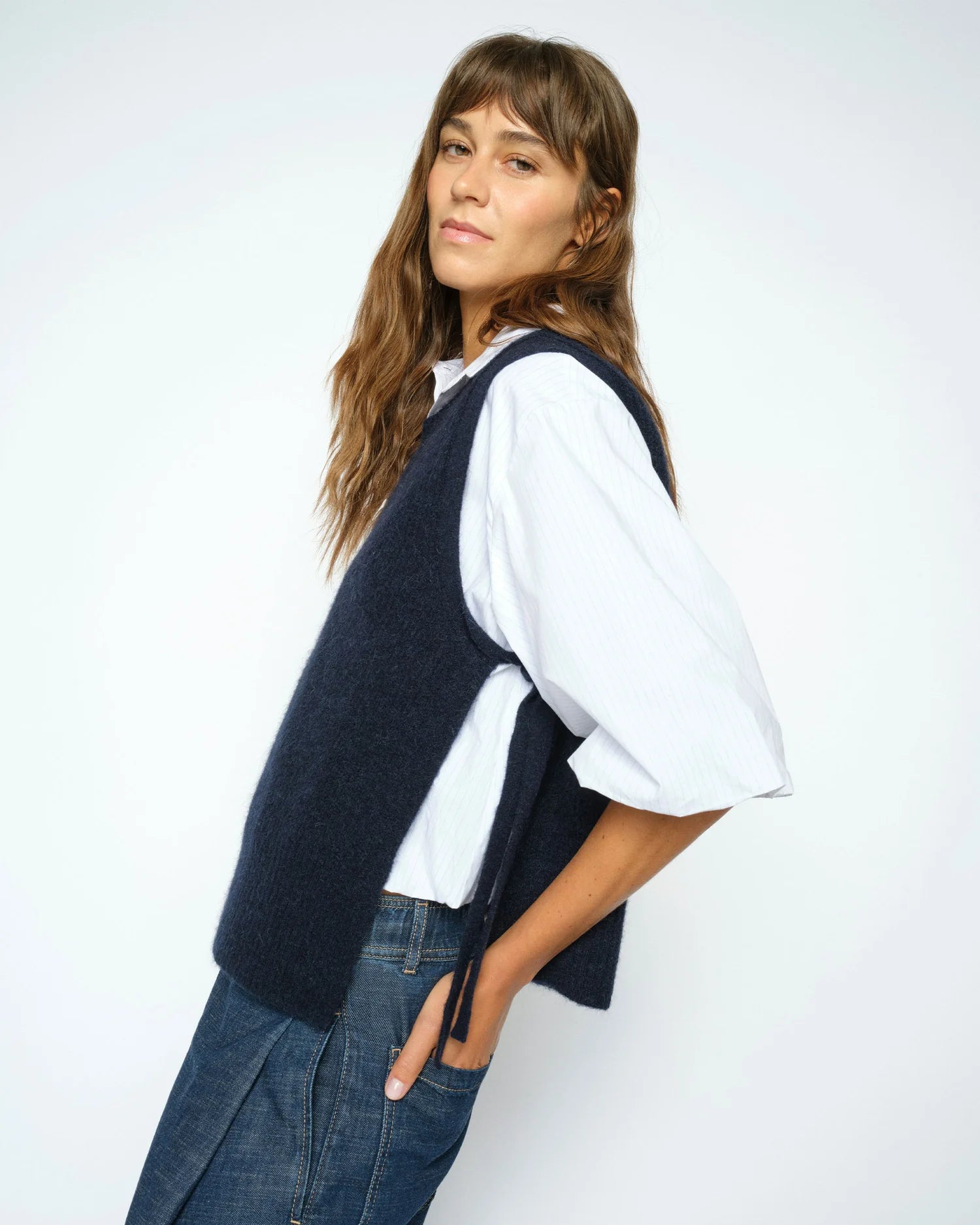 A woman with long brown hair stands sideways against a plain light background, wearing a white shirt, blue jeans, and the MOS MOSH Thora Tie Knit Vest in navy.