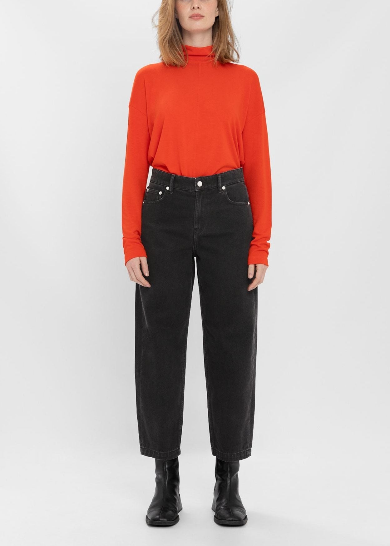 A person in a red long-sleeve top and black Navasota Denim Trousers from Loreak Mendian, featuring a high waist, stands against a plain background, while icons indicating organic cotton and commitment are visible in the top right corner.