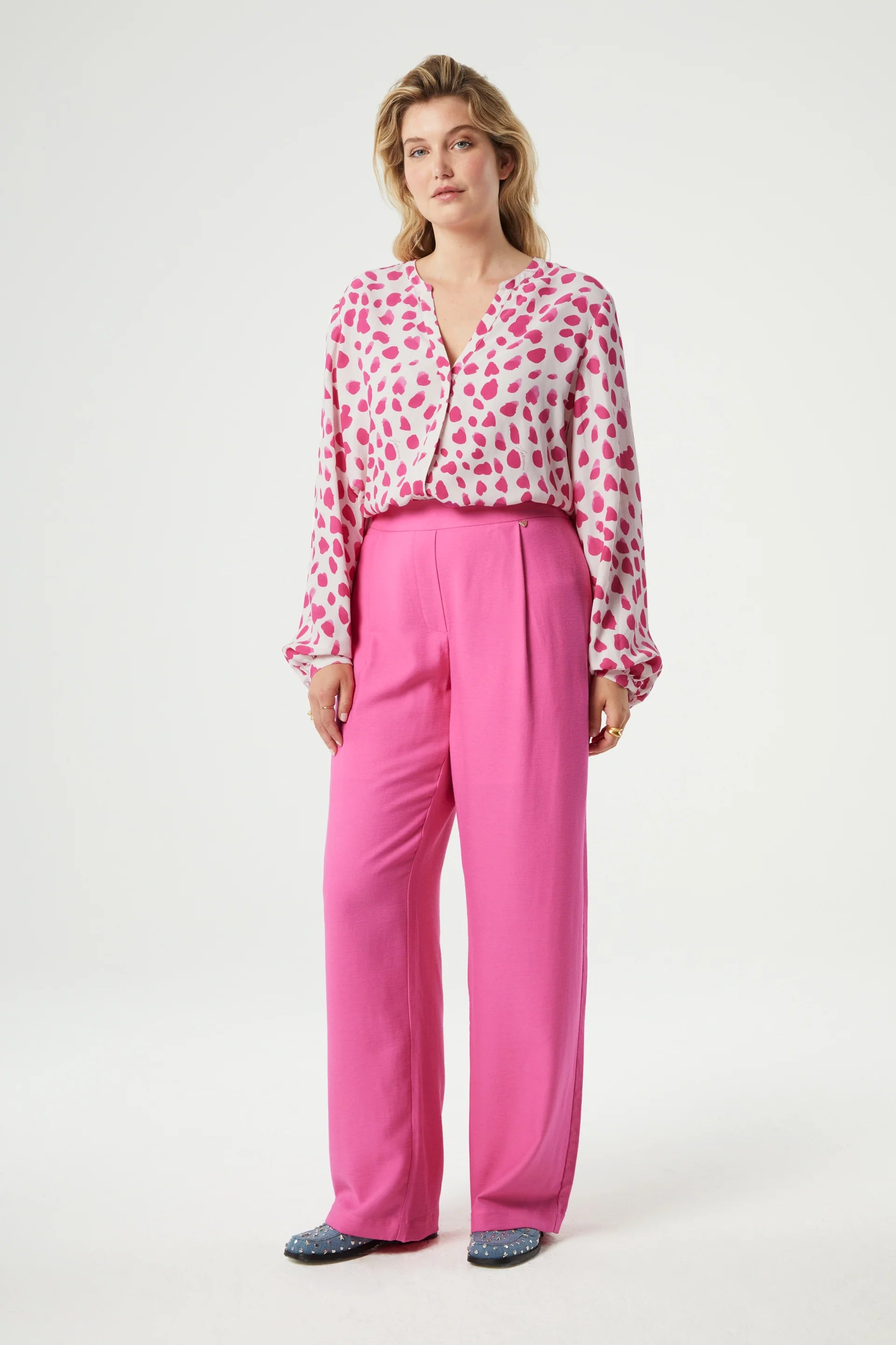 Woman wearing a pink blouse with heart patterns paired with Fabienne Chapot's Neale Trousers in Candy Pink.