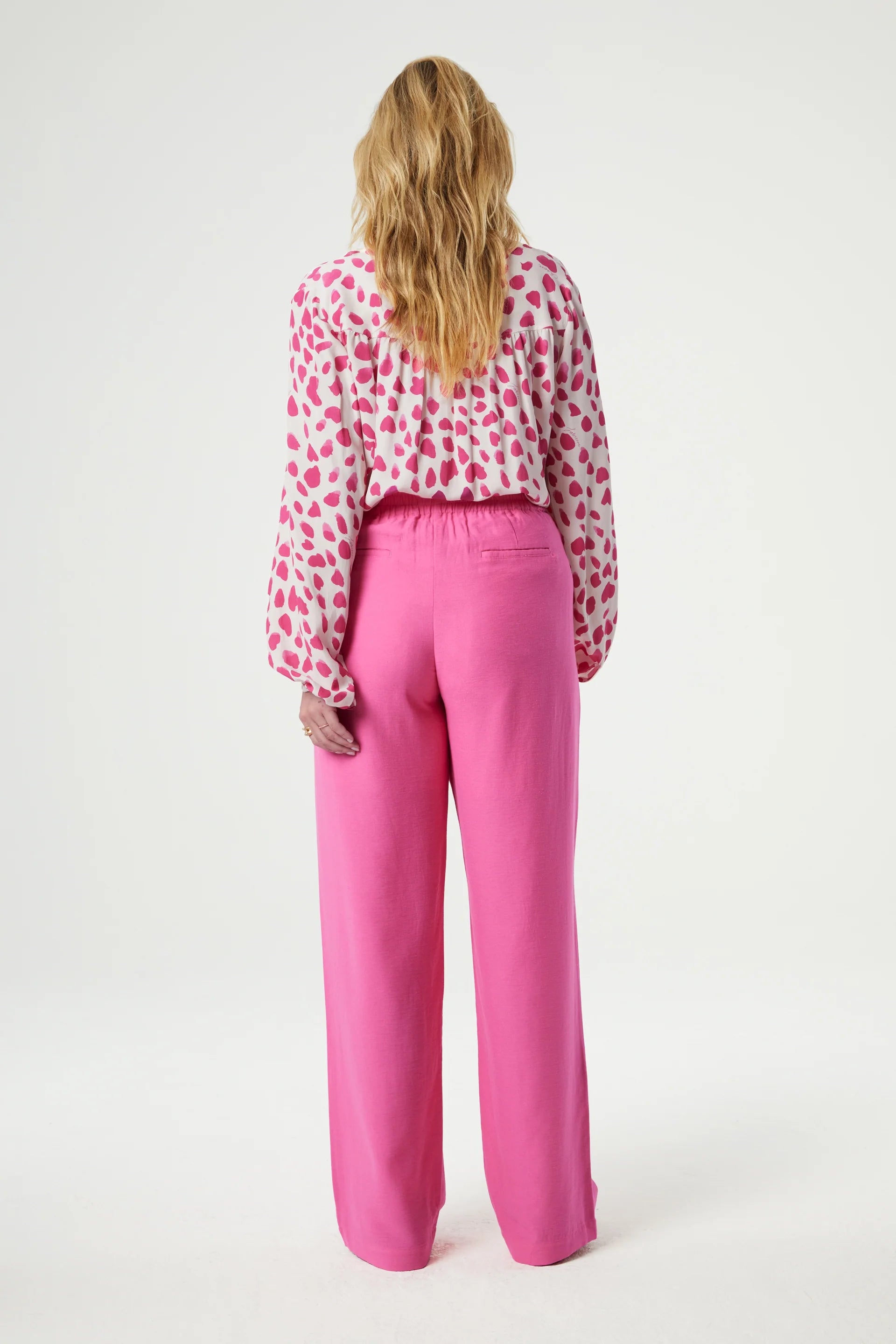 A person standing with their back to the camera, wearing a white shirt with red polka dots and Fabienne Chapot Neale Trousers in Candy Pink.