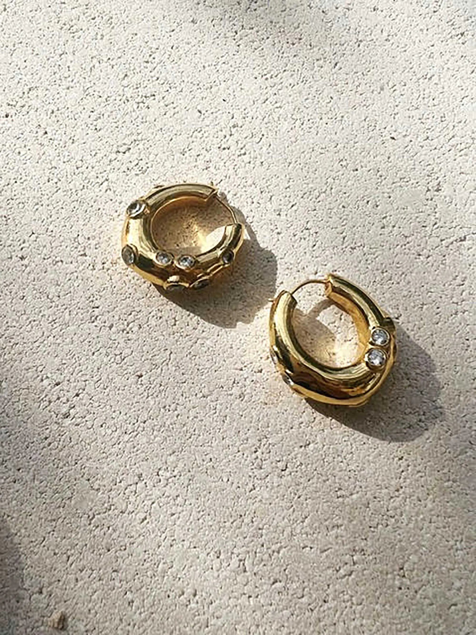 A pair of medium-sized chunky hoop earrings adorned with sparkling crystals, known as Shyla - Oren Hoops - Gold, laying on the ground.