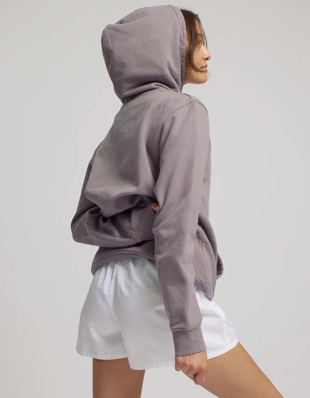 A young woman in a gray hoodie and Colorful Standard organic twill shorts is shown in profile against a neutral background.