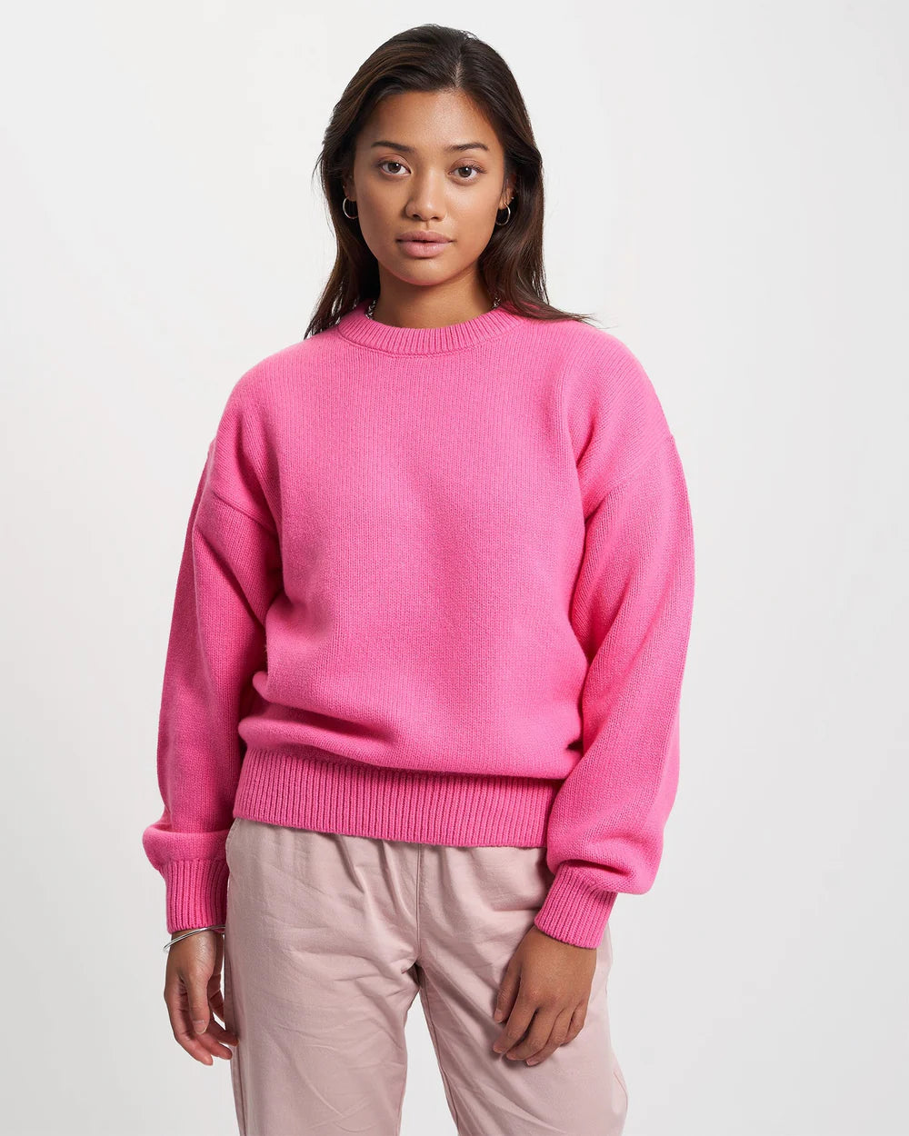 An individual is wearing the Oversized Merino Wool Crew in Magenta Blaze by Colorful Standard, paired with light pink pants, and posing against a plain white background.