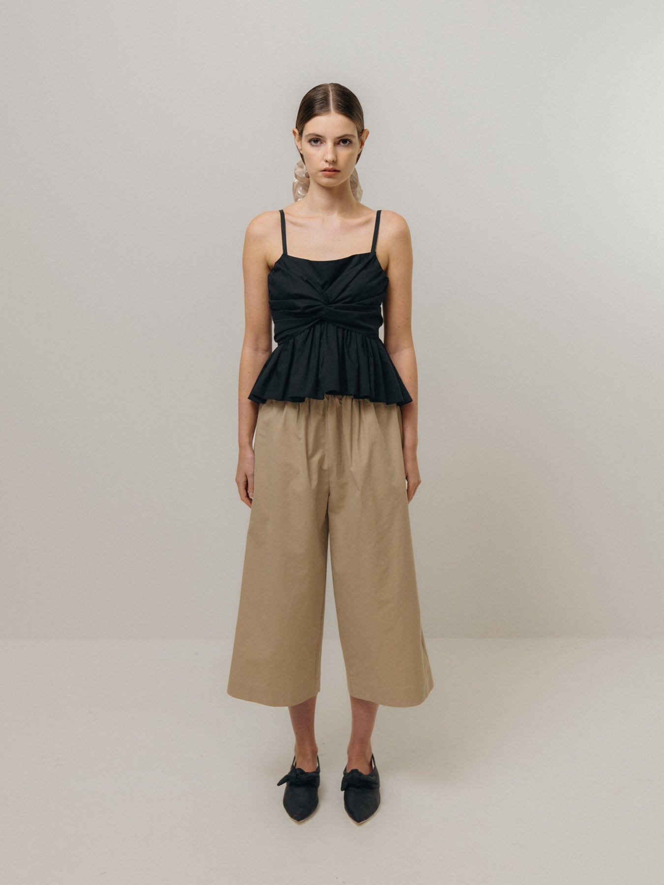 In a neutral-toned room, a woman wears an Emin and Paul black sleeveless top with beige Cotton Culottes featuring an elasticated high waist. Her black shoes complement the look, and her hair is neatly tied back.