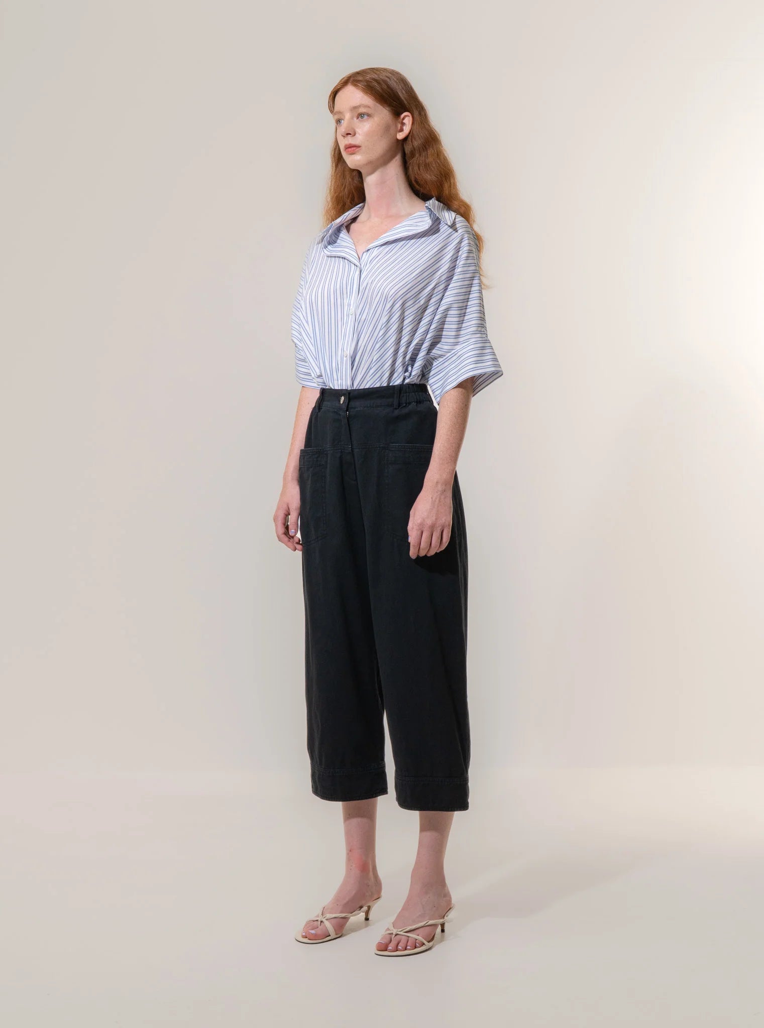 Woman standing against a plain background, wearing a loose striped shirt, Emin and Paul Utility Trousers - Black, and white heeled sandals.