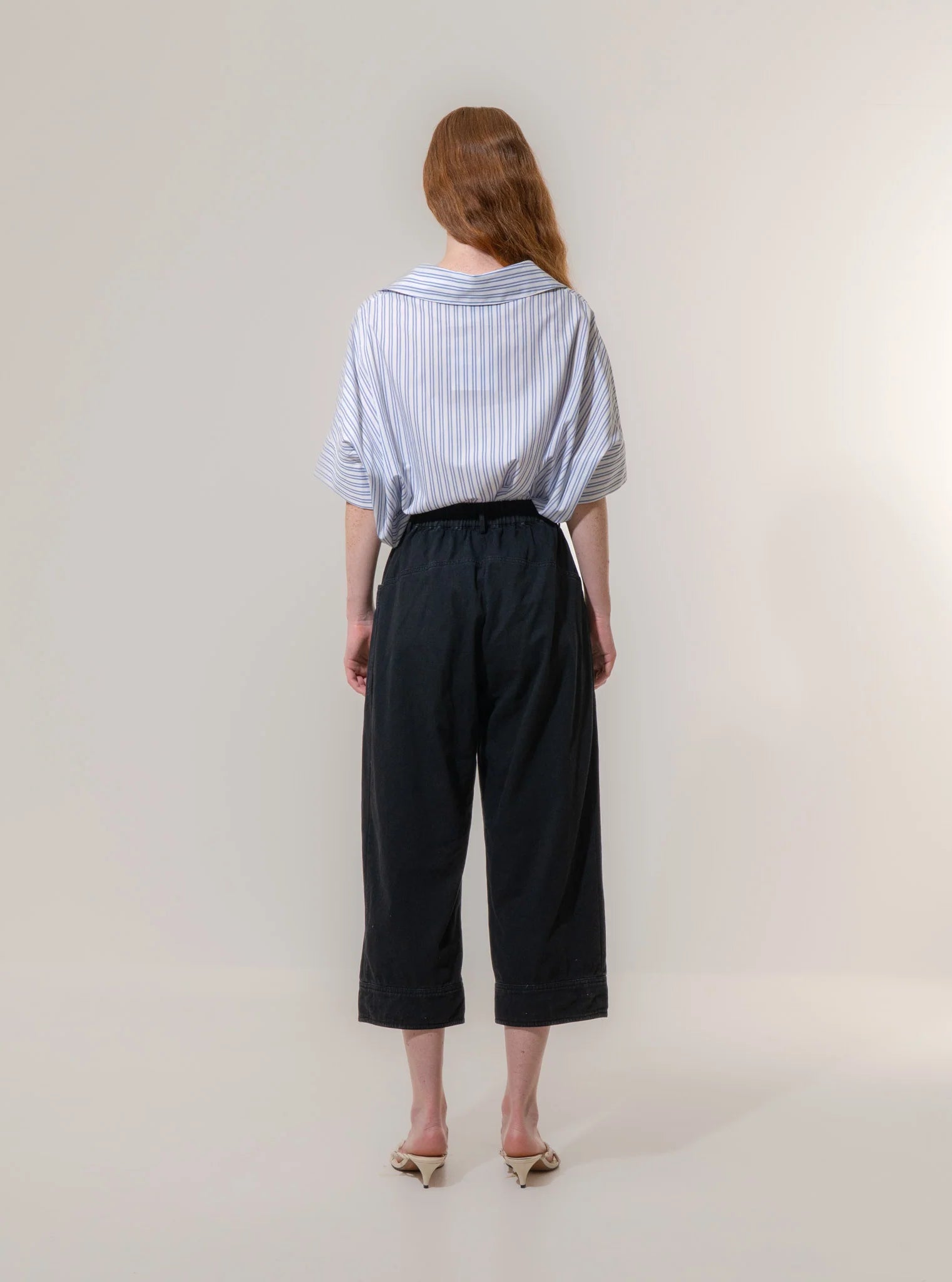 A person stands with their back to the camera, wearing Emin and Paul's Utility Trousers - Black with a loose striped shirt and beige heels, set against a plain background.