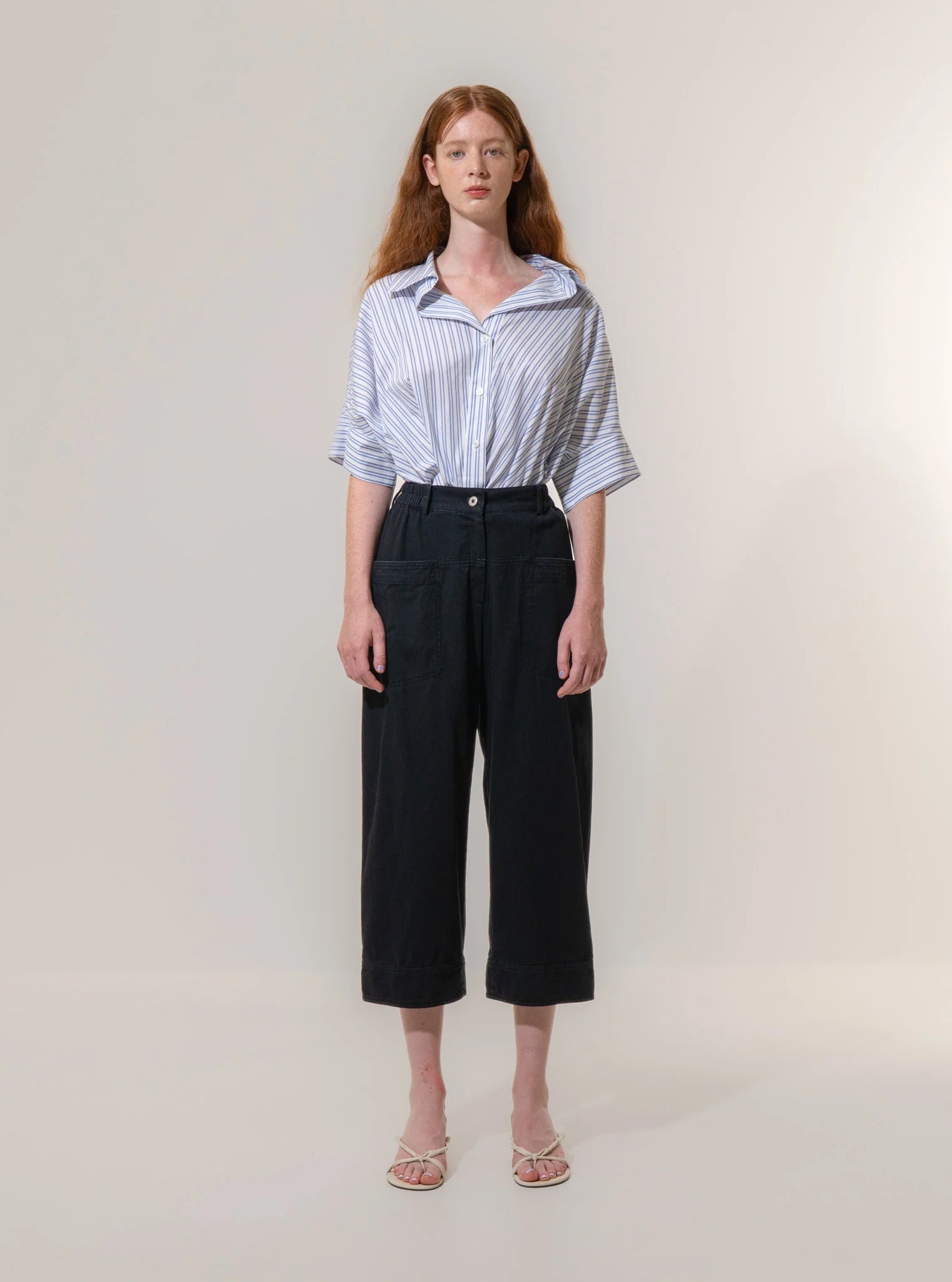 A person stands against a plain background wearing a blue and white striped shirt, Emin and Paul Utility Trousers - Black, and white sandals.