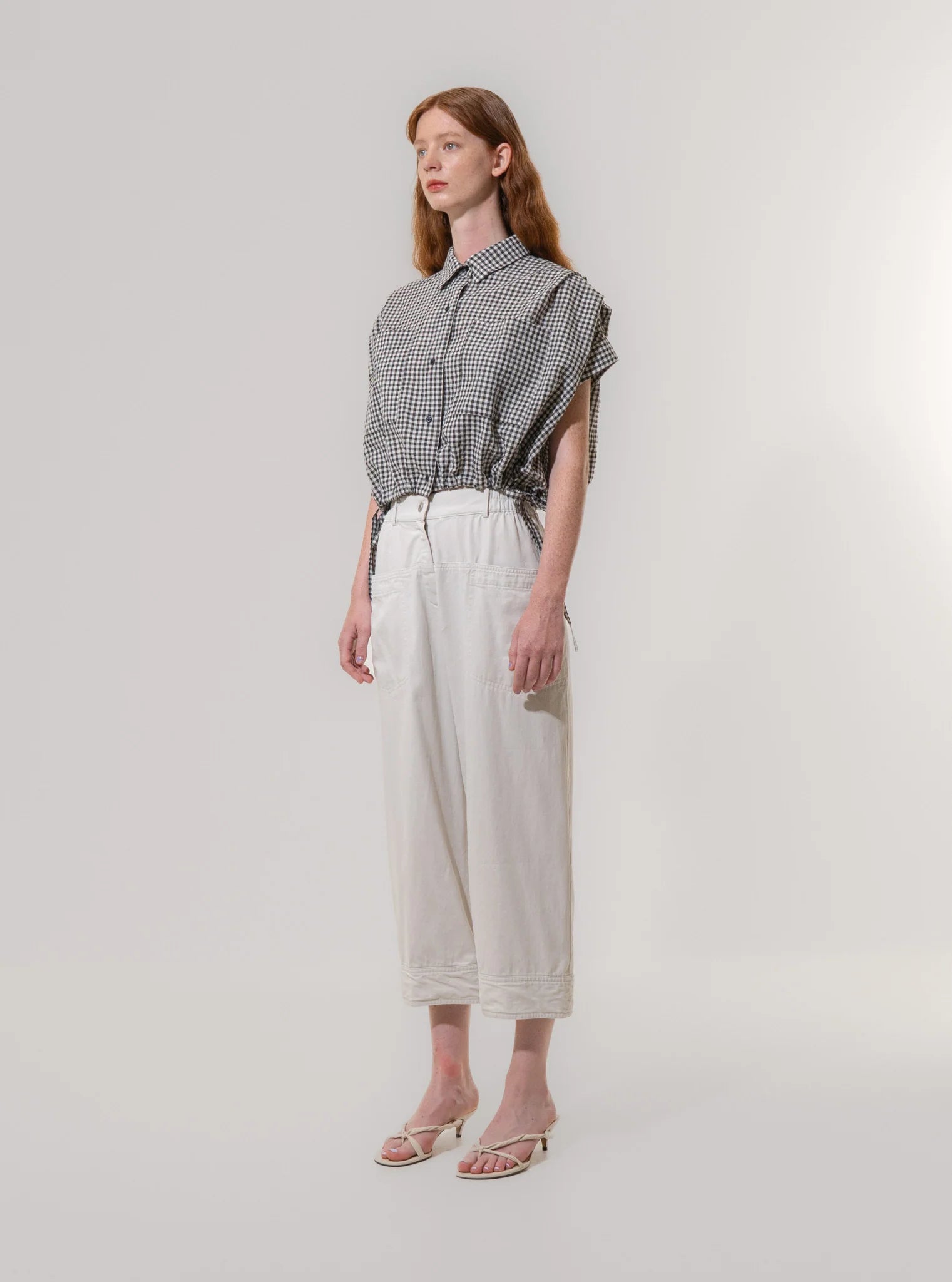 A person stands against a plain background wearing a black-and-white checkered short-sleeve shirt, Emin and Paul Utility Trousers - Light Grey (100% cotton), and white sandals.