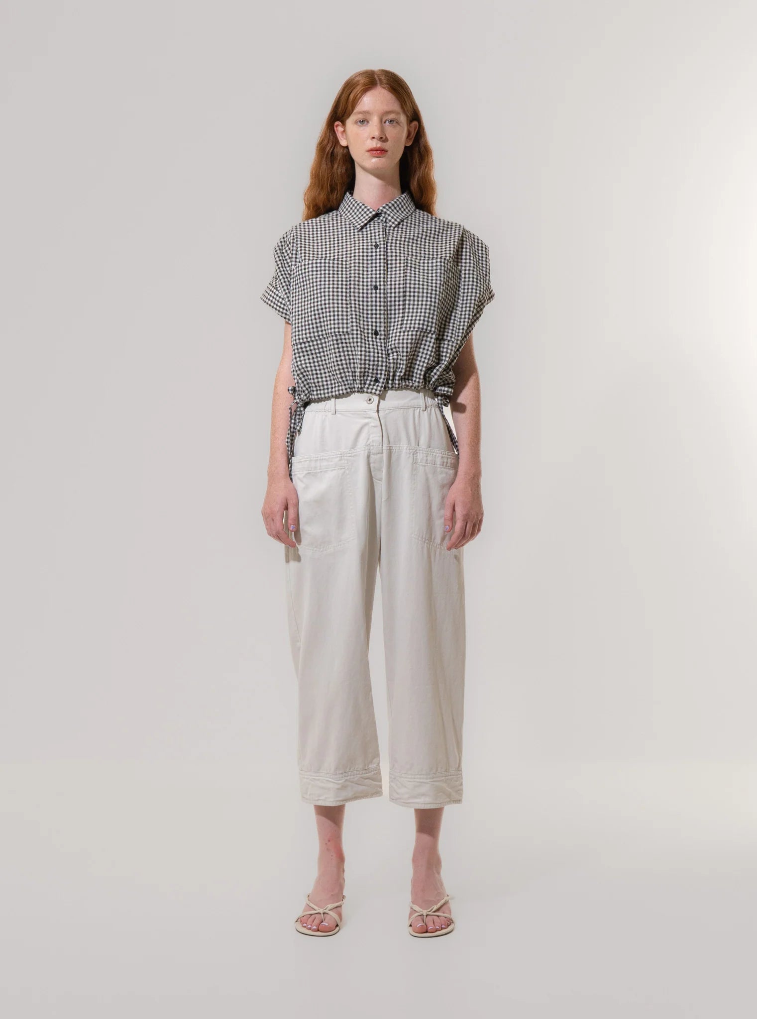 A person stands facing forward against a plain background, wearing a black and white checkered short-sleeve shirt, Emin and Paul Utility Trousers - Light Grey (100% cotton), and white sandals.