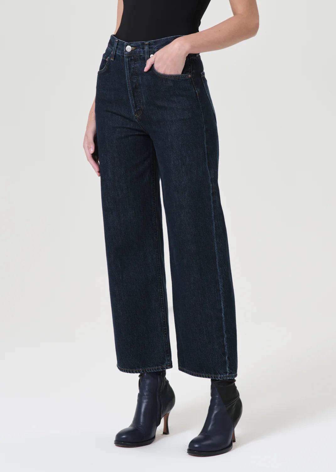 A person wearing AGOLDE's Ren High Rise Wide Leg Jeans in Polished vintage indigo denim, a black top, and black heeled boots stands against a plain background.