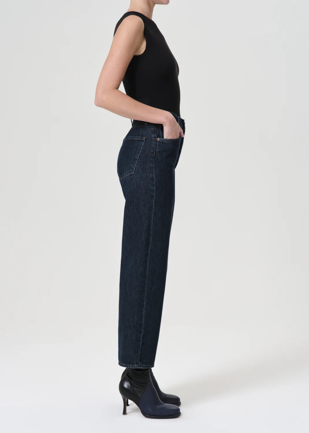 A person stands in profile wearing a black sleeveless top, AGOLDE's Ren High Rise Wide Leg Jeans - Polished in vintage indigo denim, and black heeled boots, against a plain white background.