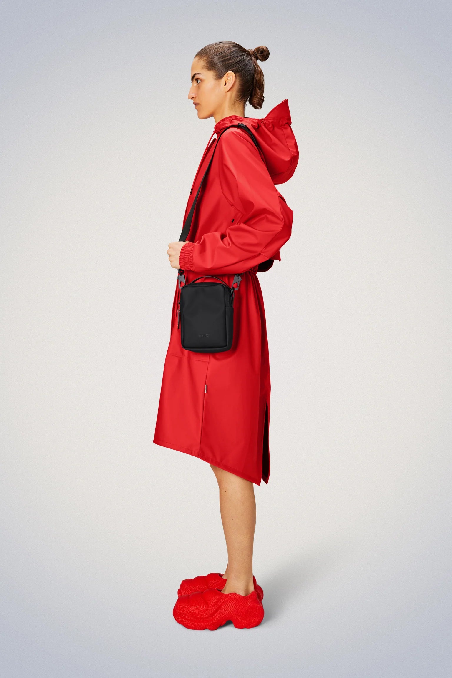 A woman, donning a red raincoat made from Rains' signature waterproof PU fabric, walks confidently with her matching Rains Reporter Box Bag.